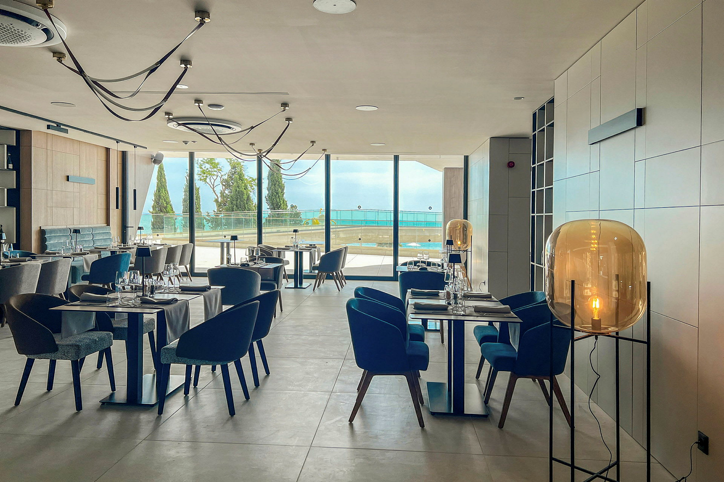 Restaurantinteriør med moderne design, oppstilte bord og stoler og store vinduer med utsikt over havet. (AI-generert tekst)