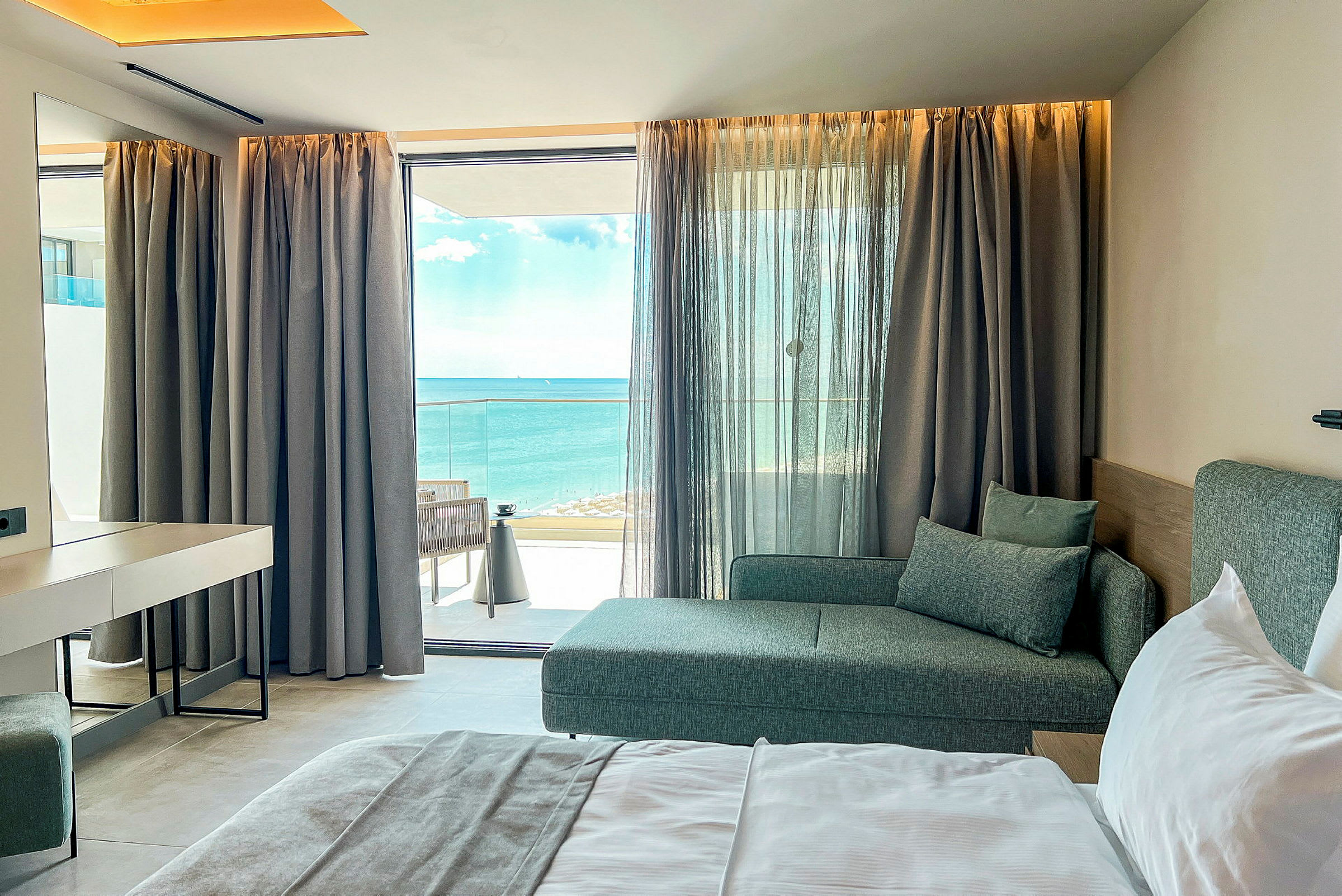 Hotellrom med seng, sofa og havutsikt gjennom en vindusfront. (AI-generert tekst)