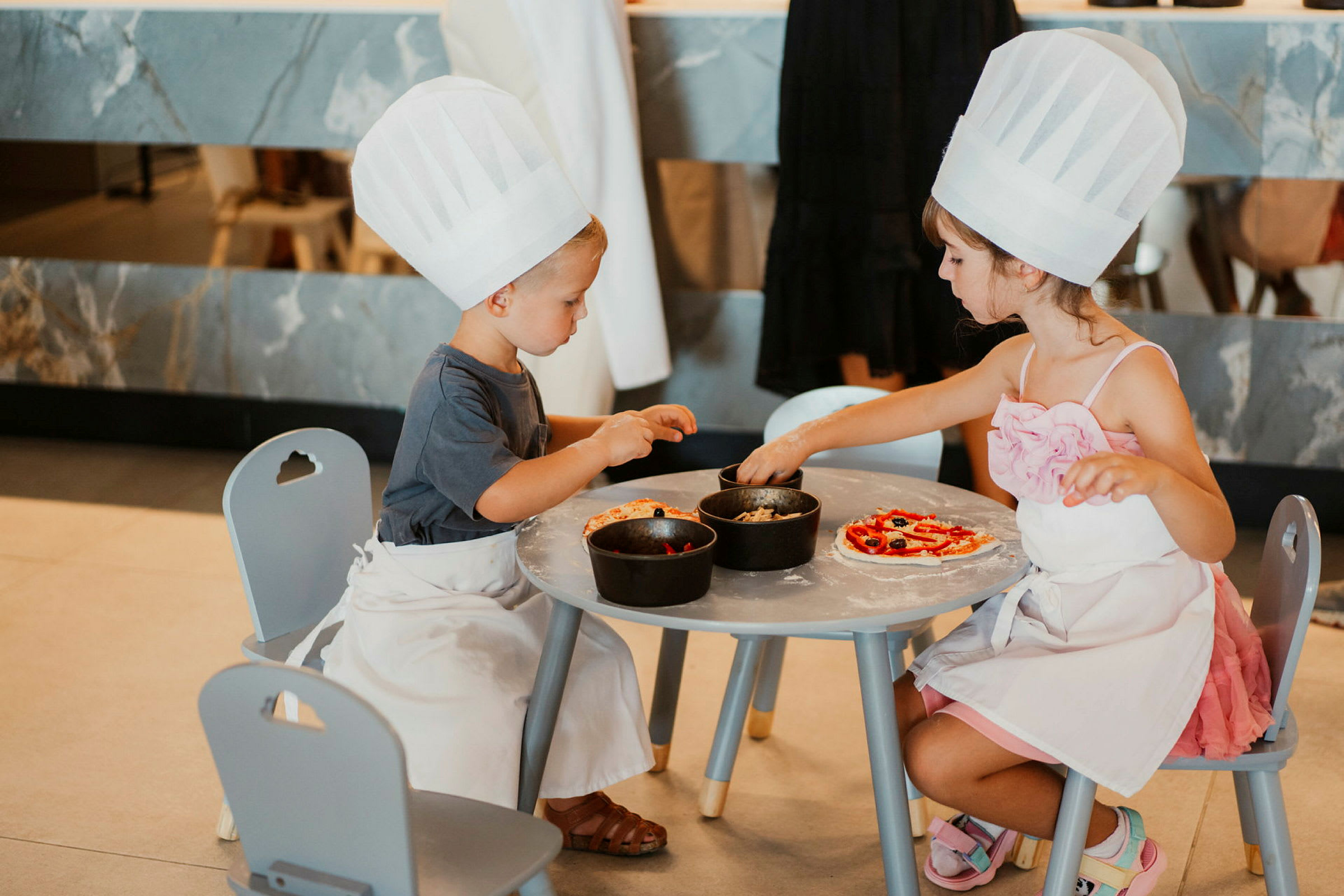 To barn med kokkehatter og forklær som dekorerer pizzaer ved et bord. (AI-generert tekst)