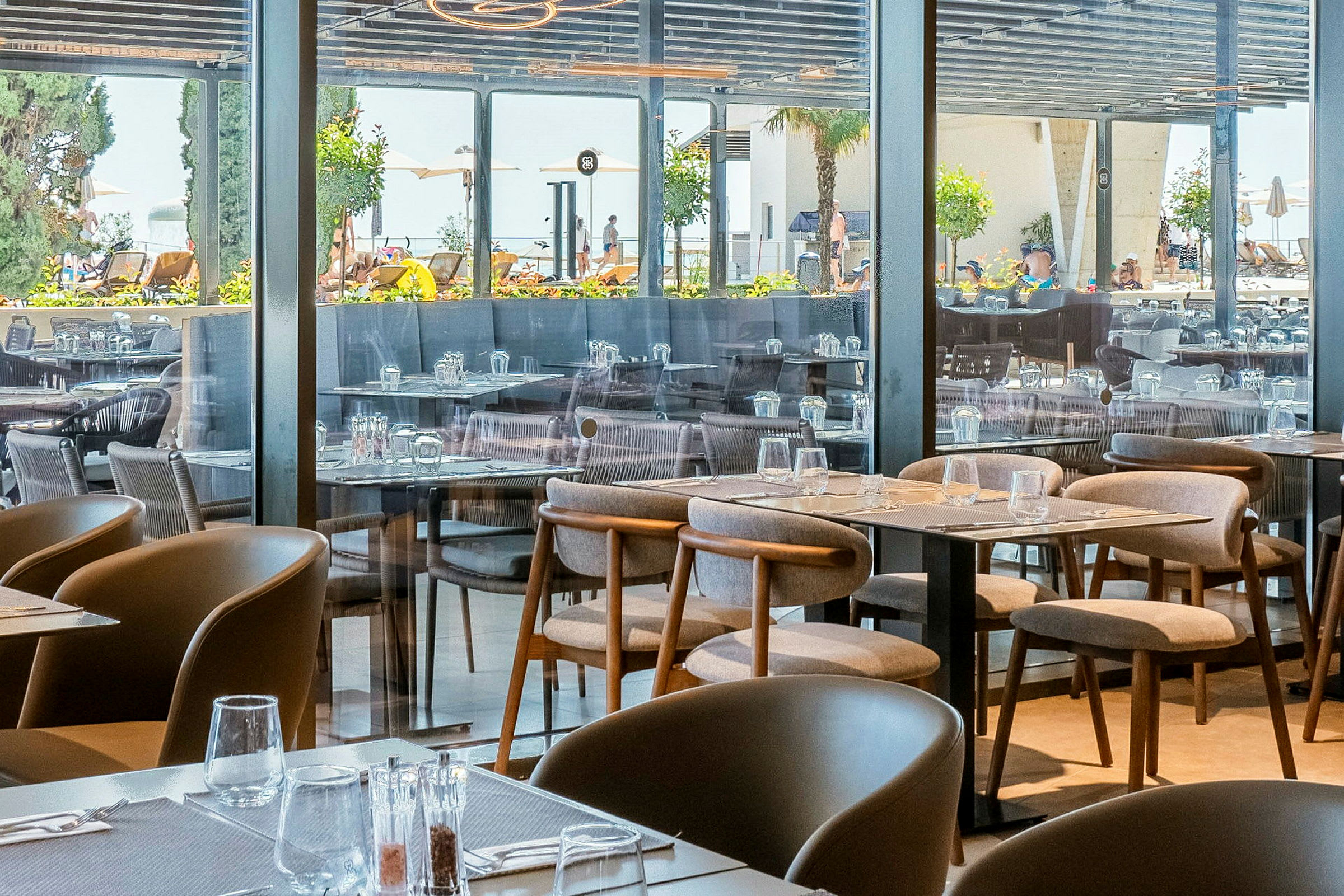 Bilde av en restaurant med oppstilte bord og stoler, med utsikt mot utendørsområdet gjennom glassvegger. (AI-generert tekst)