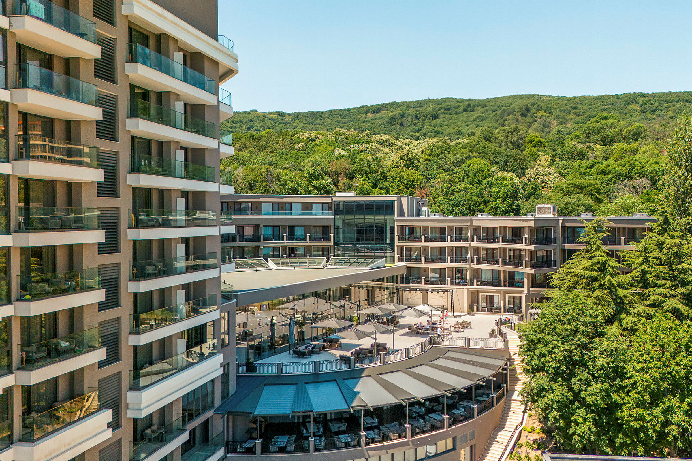 Flere hotellbygninger omgitt av grønne områder og en terrasse med parasoller. (AI-generert tekst)