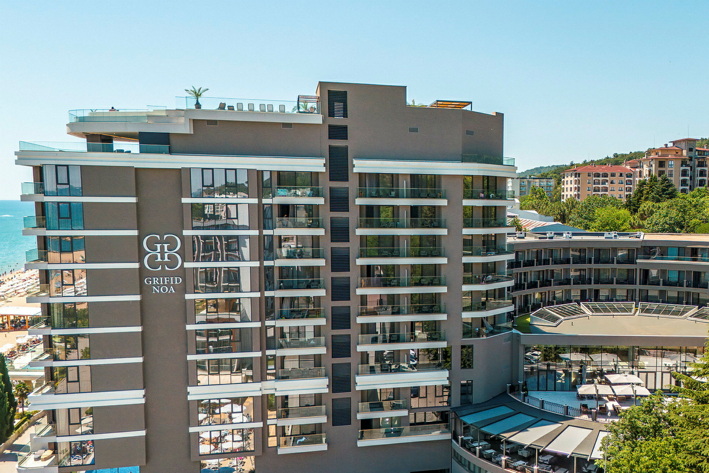 Moderne hotell med flere etasjer, stor logo på fasaden, beliggende nær en strand og omgitt av bygninger. (AI-generert tekst)