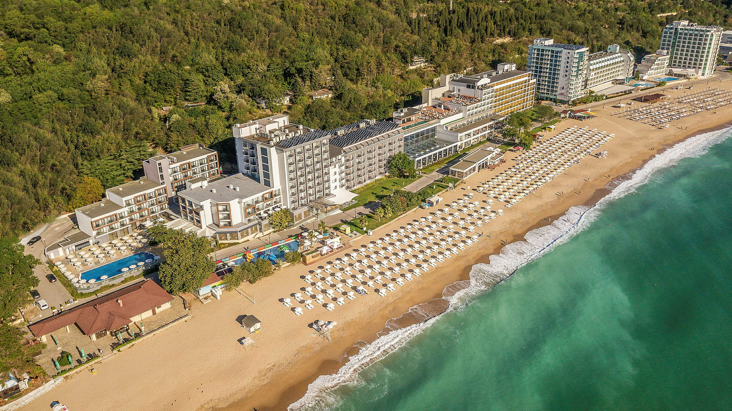 Utsikt over hotell ved kysten, med strand og parasoller samt grøntområder i bakgrunnen. (AI-generert tekst)