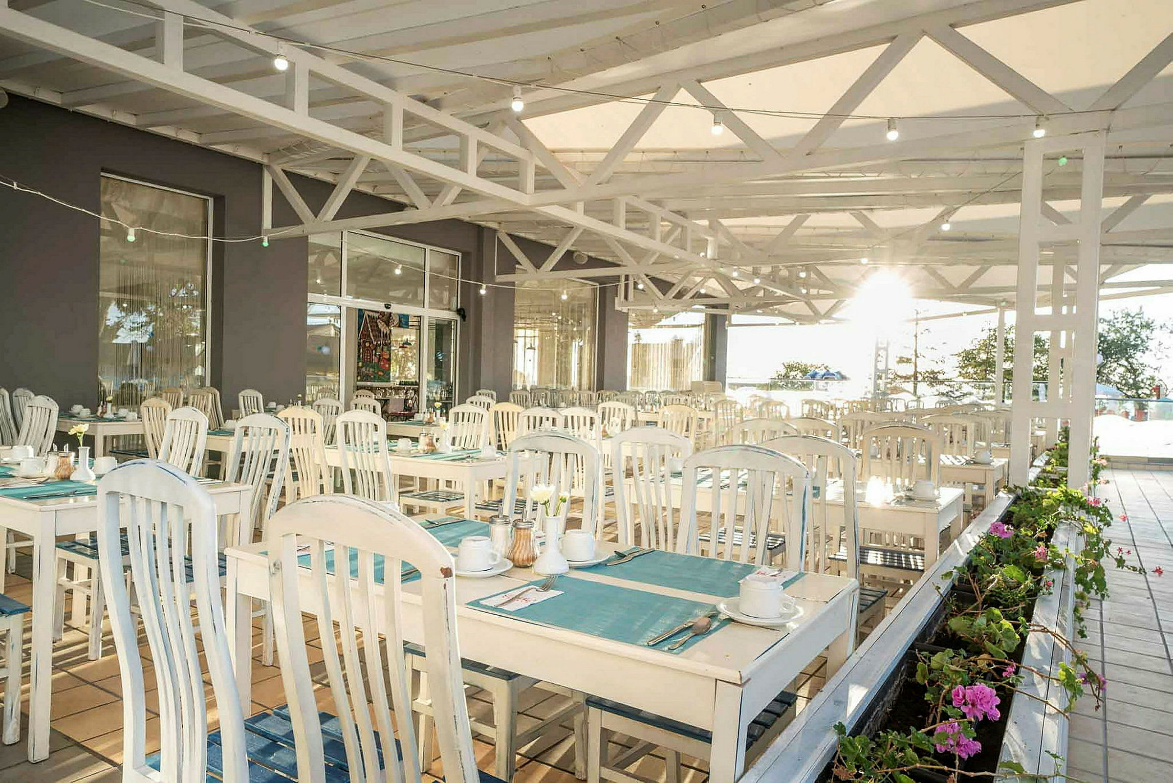 En utendørs restaurant med bord og stoler i lys trestil, dekket med tallerkener og kopper, samt beplantning i forgrunnen. (AI-generert tekst)