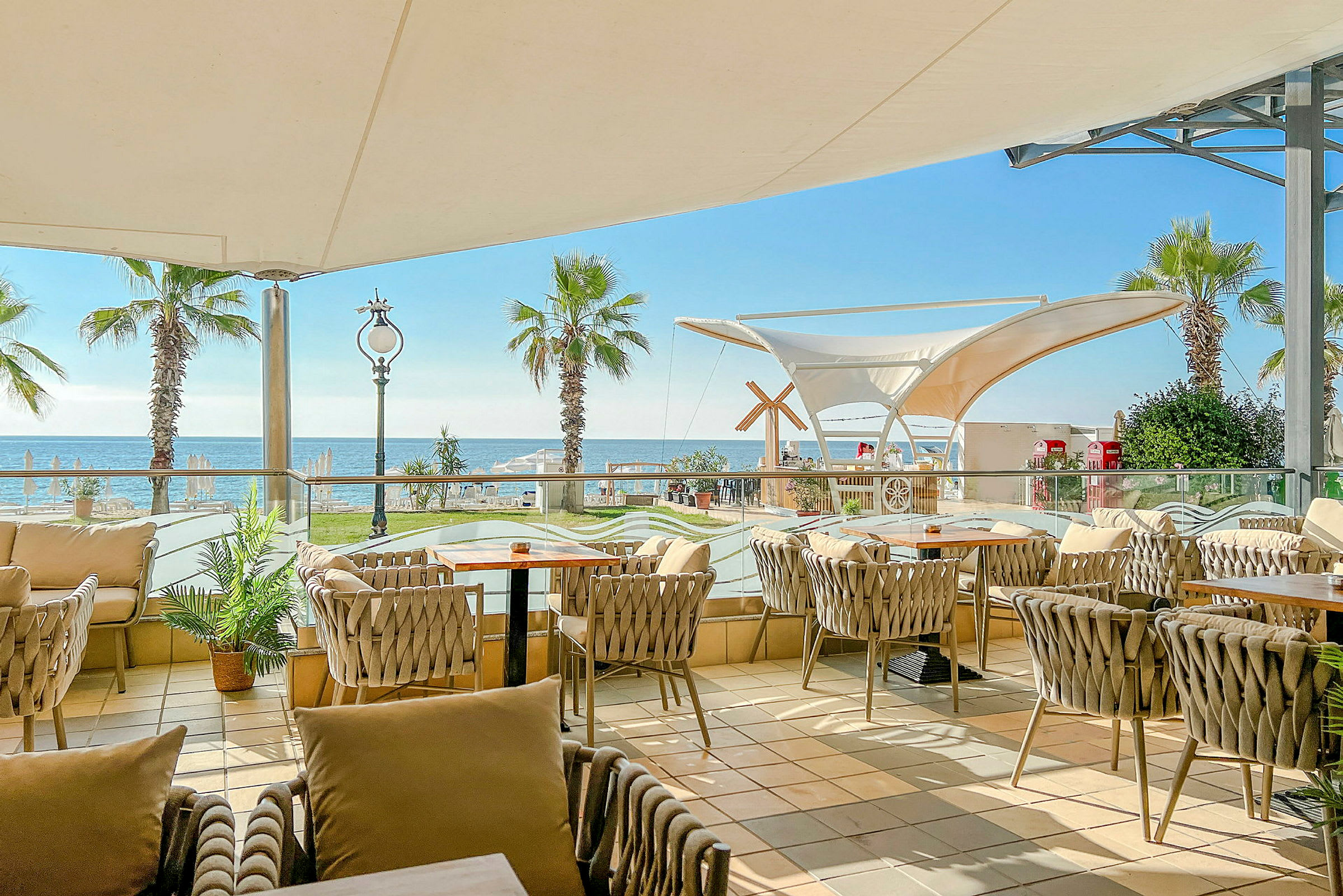 En terrasse med bord og stoler, utsikt mot havet og palmer. (AI-generert tekst)
