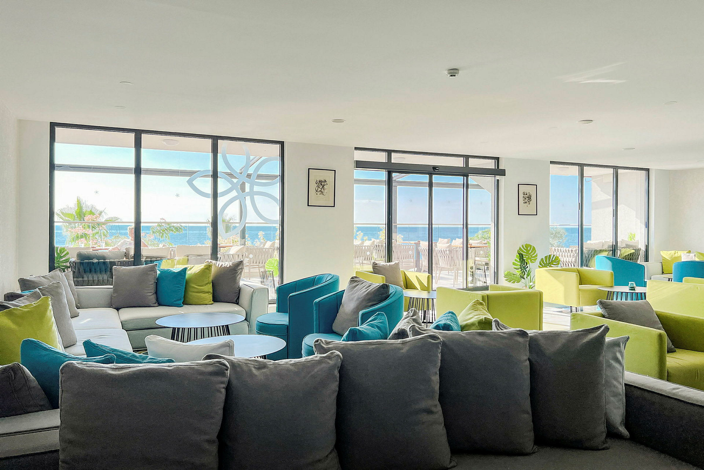 En lounge med sofaer og fargerike stoler, og vinduer som vender mot en terrasse og havet. (AI-generert tekst)