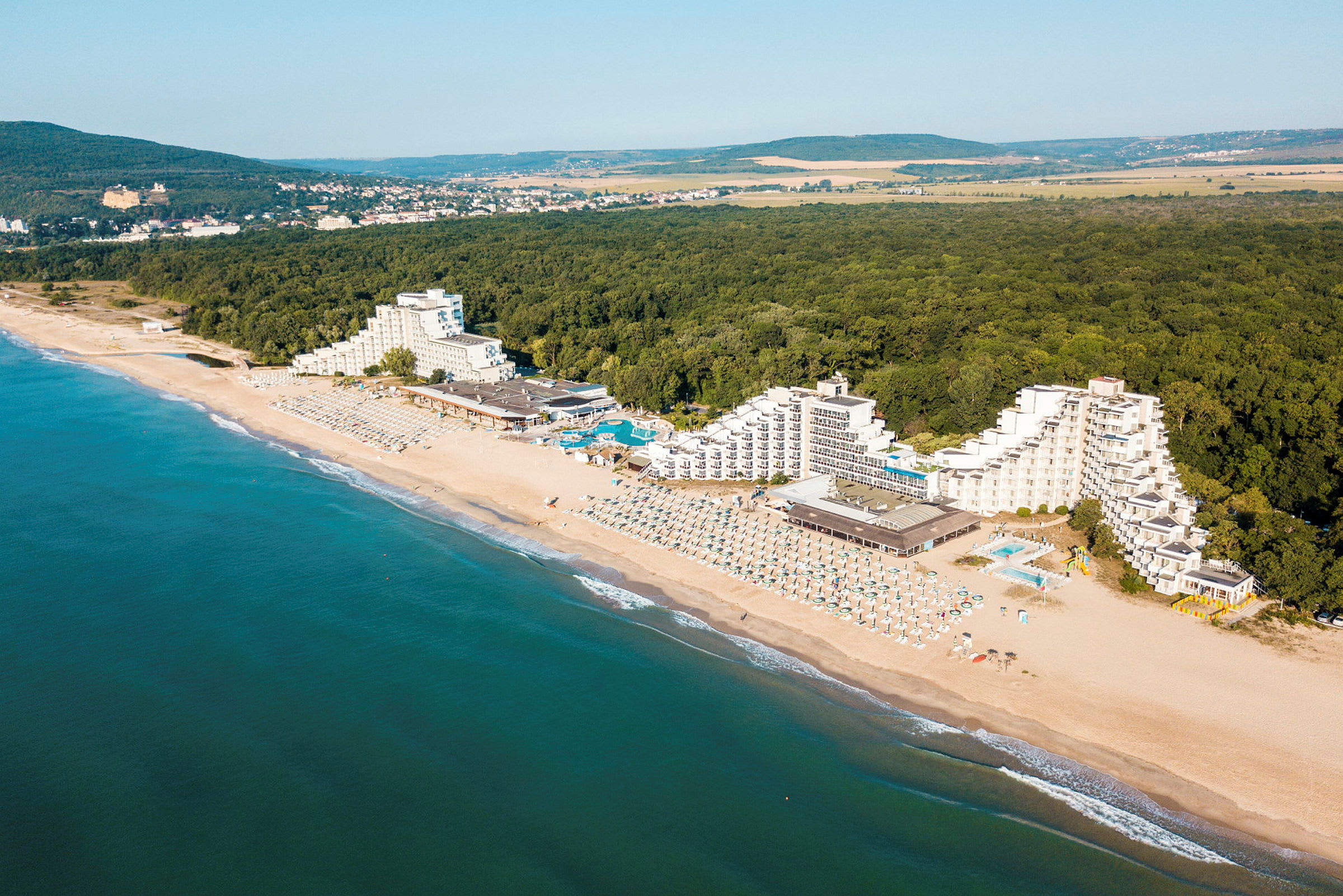 Luftperspektiv av hotell ved en strand med grønne skoger i bakgrunnen. (AI-generert tekst)