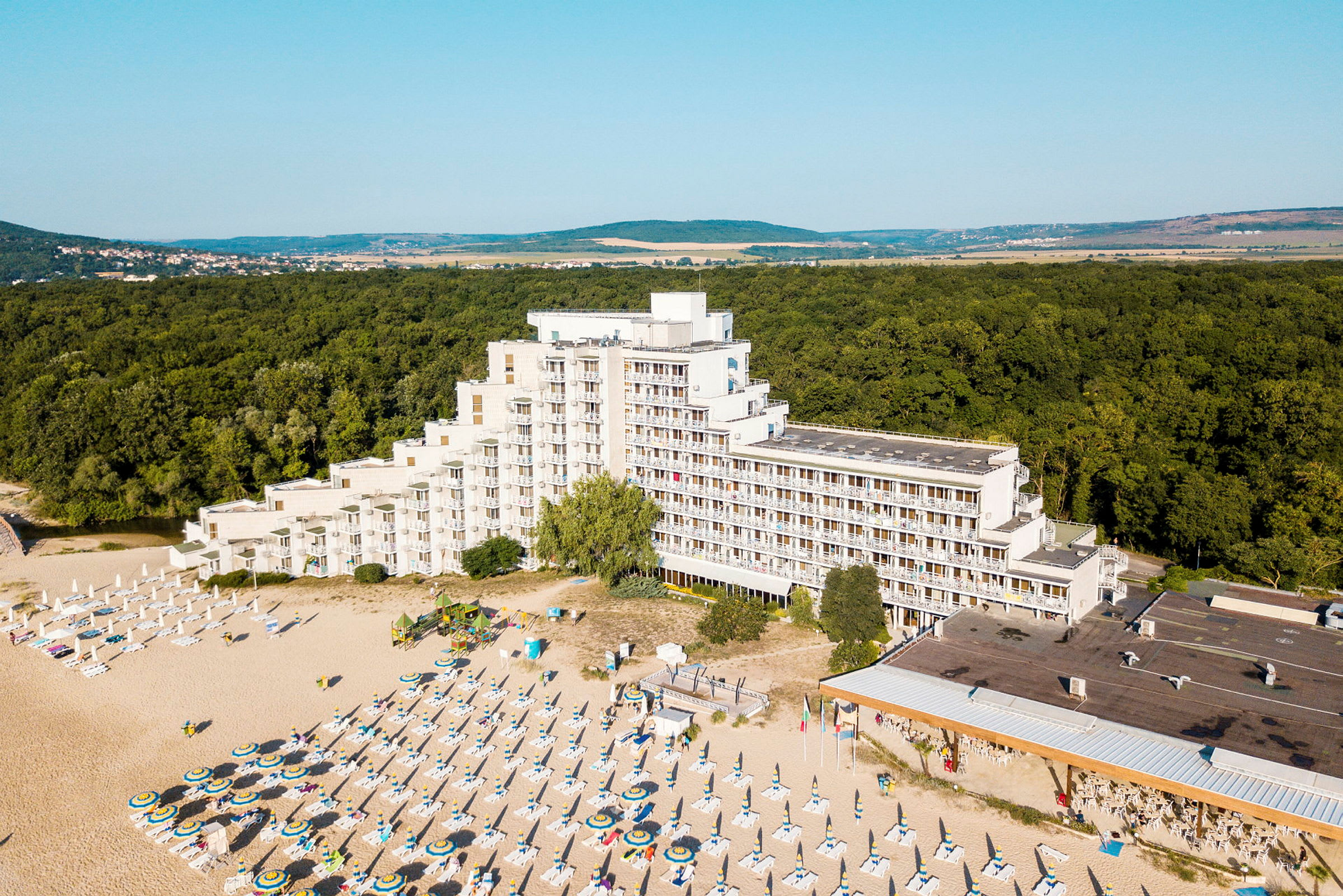 Bilde av et hotell ved en strand med solsenger og parasoller, omgitt av skog. (AI-generert tekst)