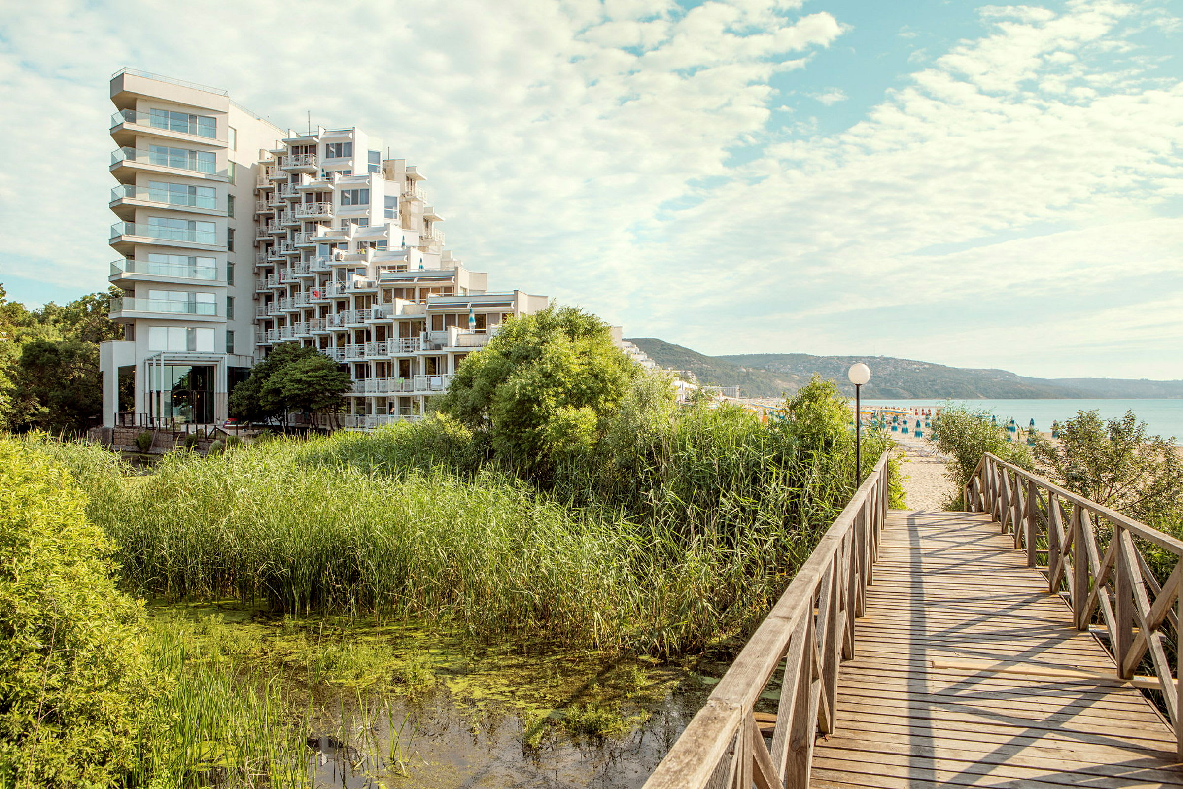 En moderne hotellbygning ligger nær et område med grønne omgivelser og vann, og en trebro leder mot en sandstrand. (AI-generert tekst)