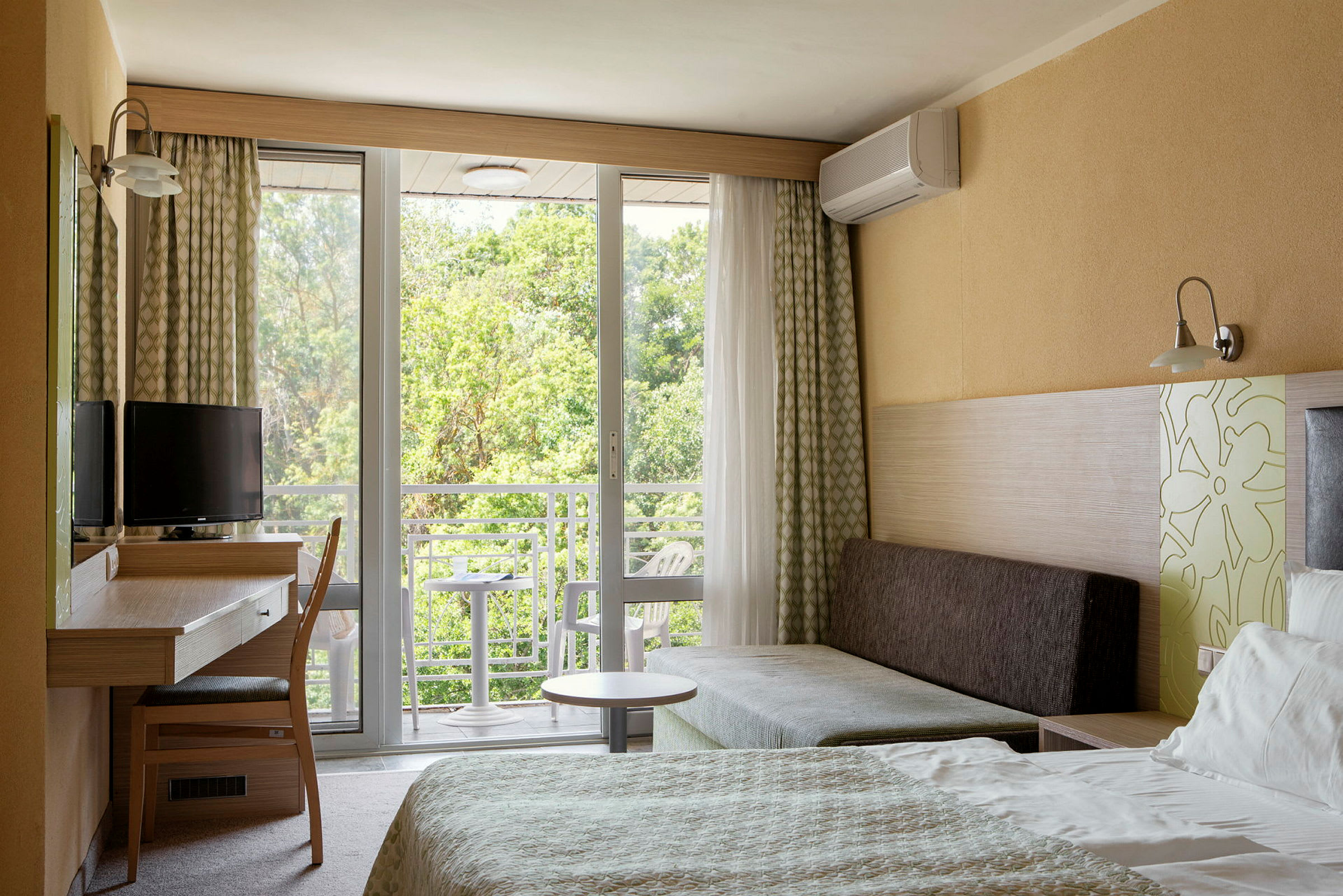 Et hotellrom med seng, skrivebord, stol og sofa. Vindu og balkong med utsikt mot grøntområder. (AI-generert tekst)