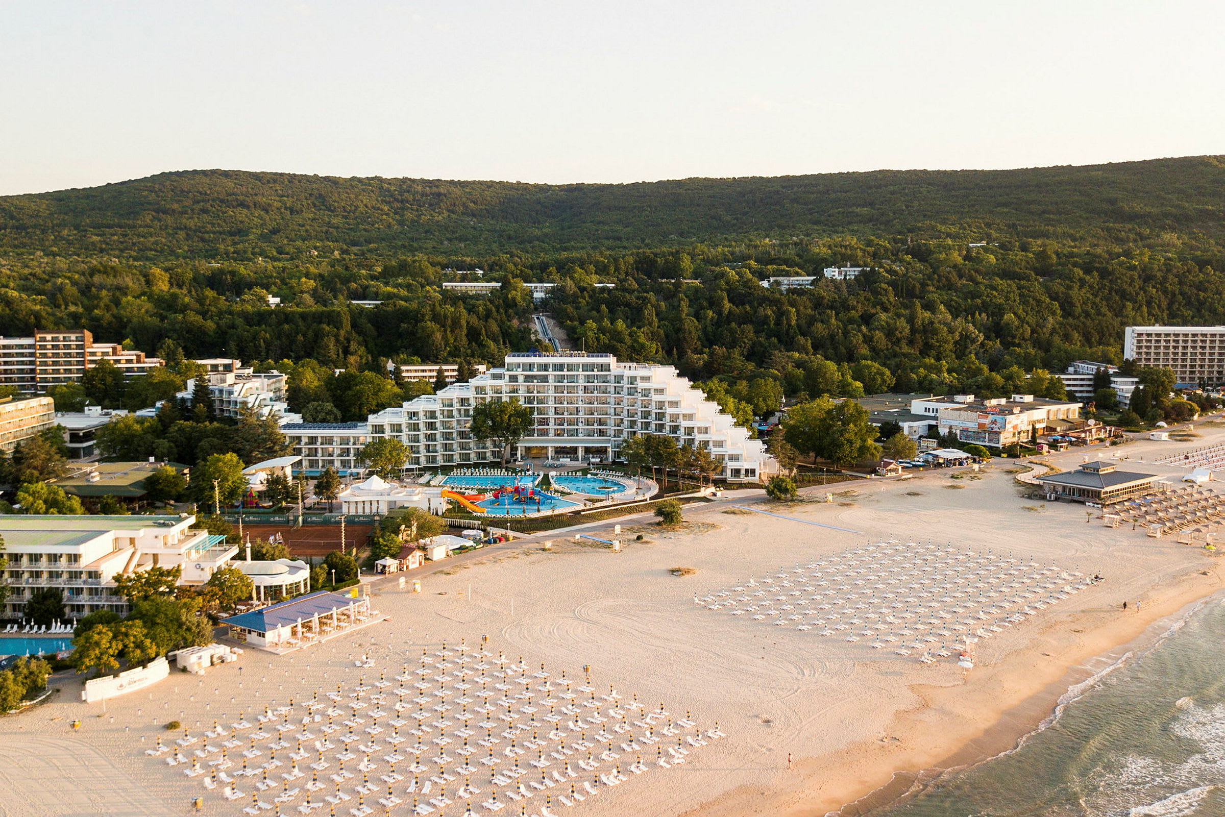 Bilde av en kystnær ferieby med hotell, bassengområde og strand med parasoller. (AI-generert tekst)