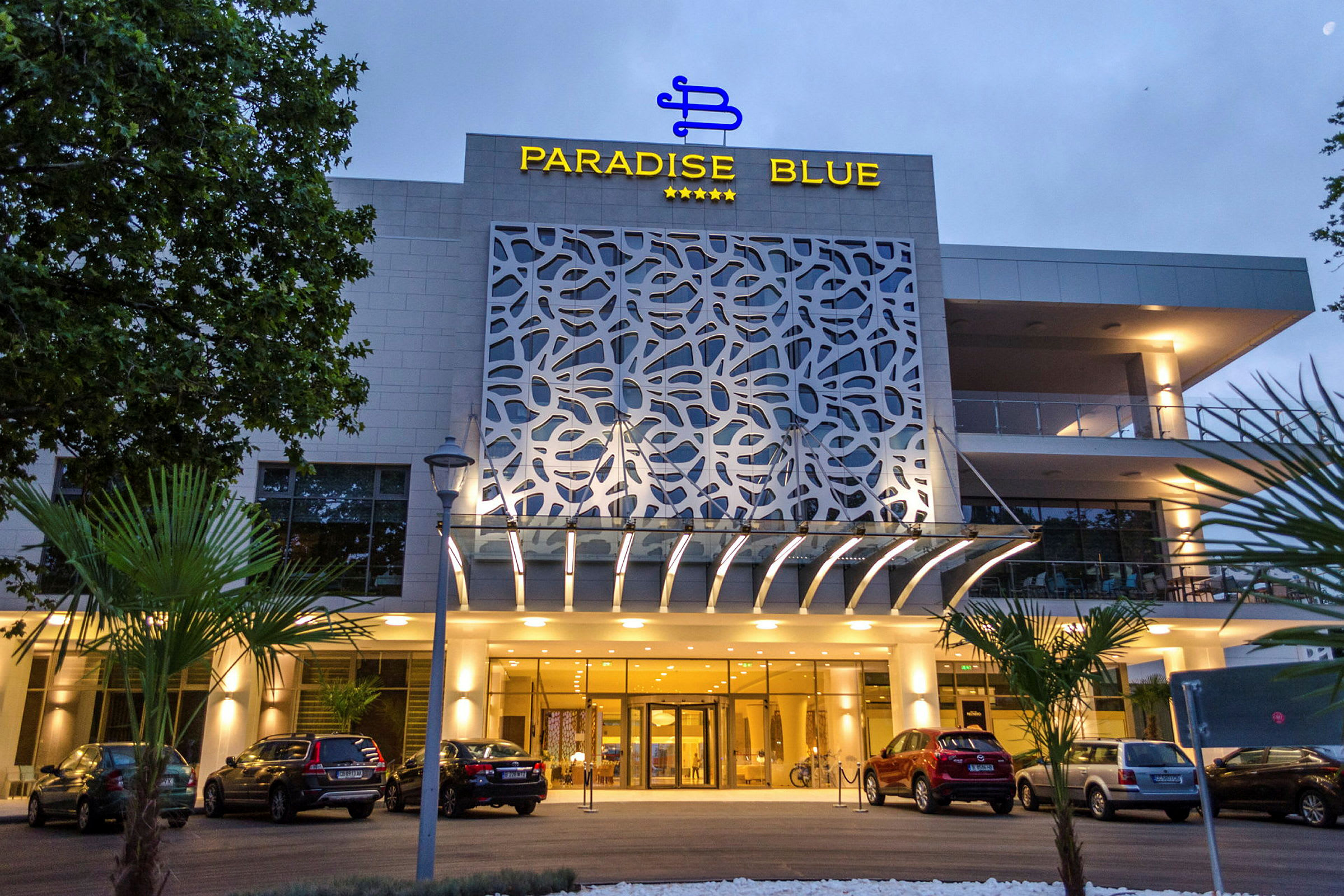 Fasaden til Paradise Blue hotell med bilparkering og inngang. (AI-generert tekst)