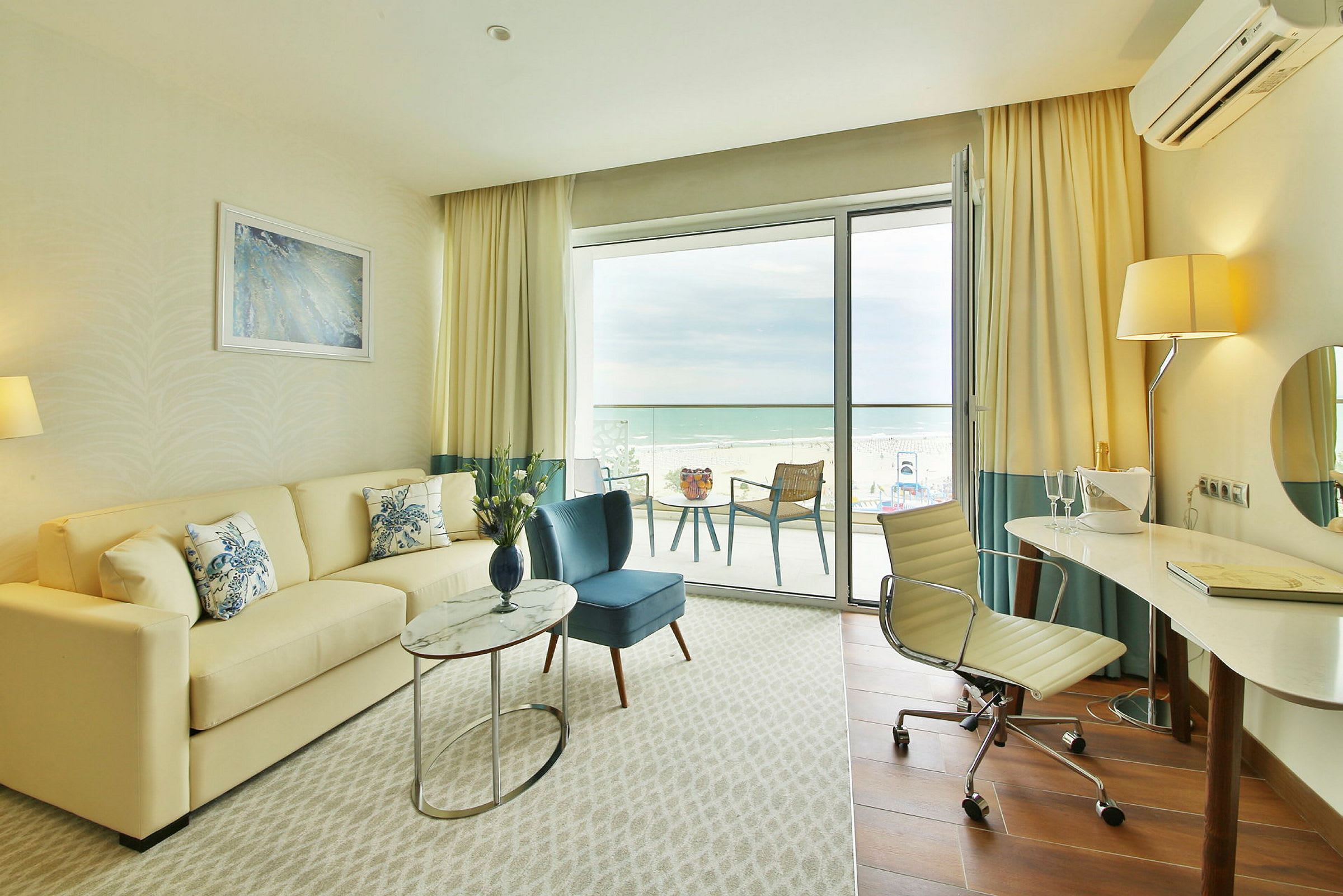 Et lyst hotellrom med en sofa, lenestol, et skrivebord og en balkong med utsikt over stranden. (AI-generert tekst)
