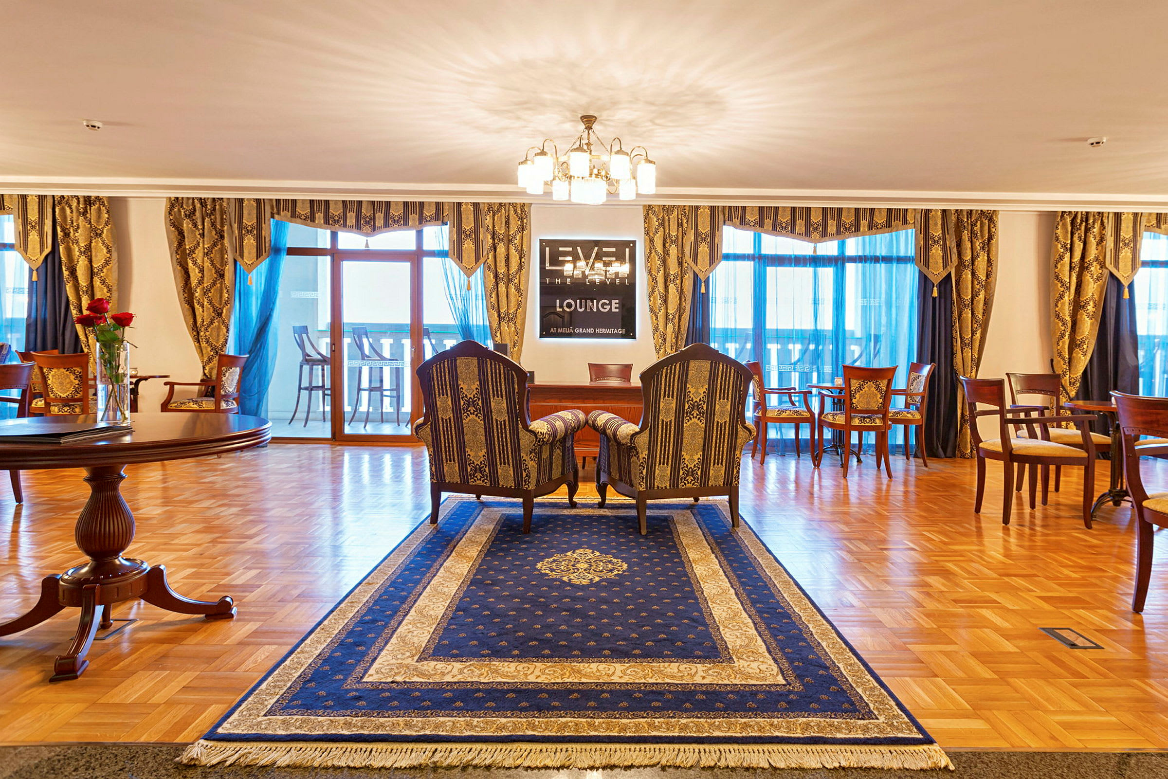 En godt innredet lounge med et blått teppe, par stoler og et bord, lamper og dekorative gardiner. (AI-generert tekst)