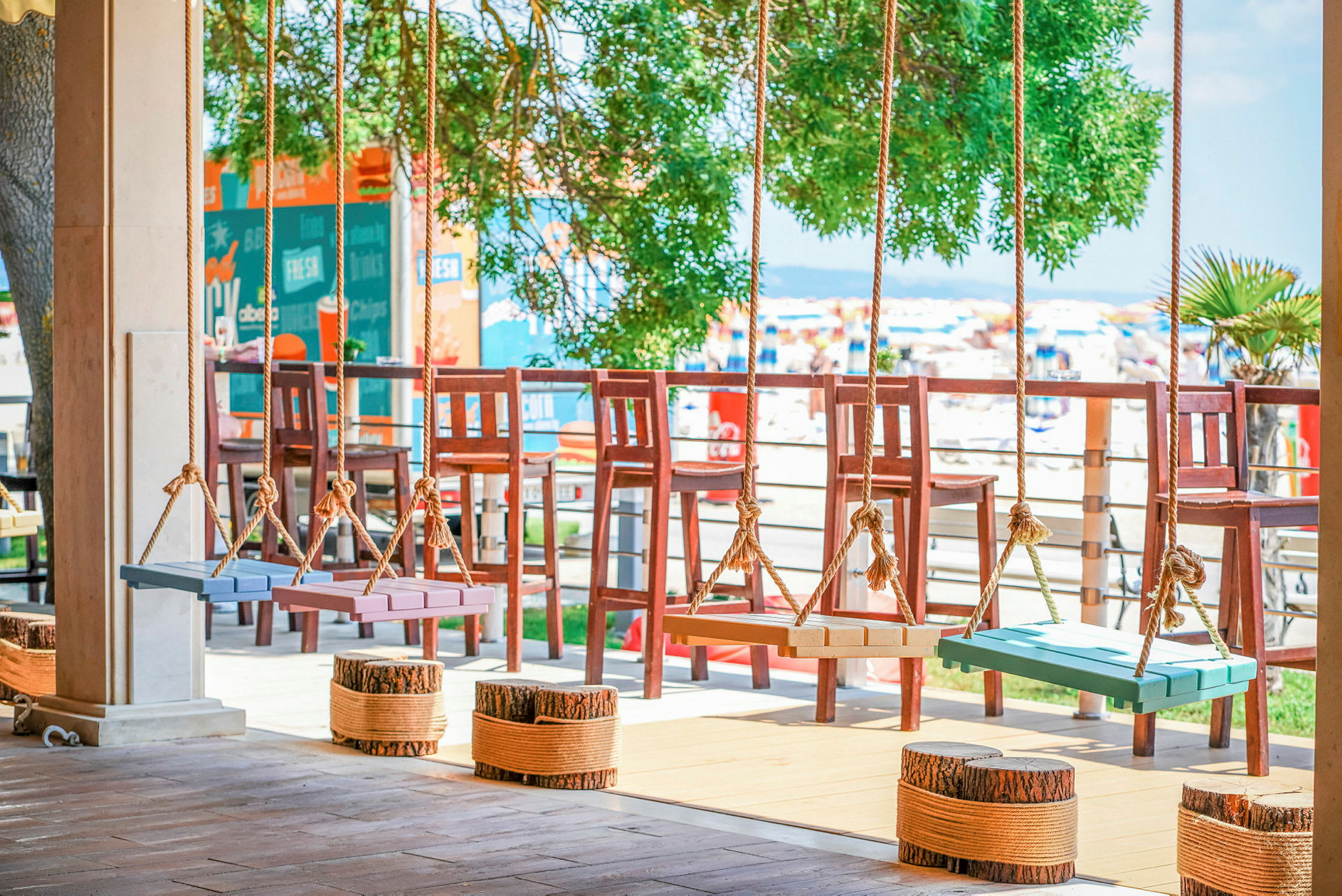 Fargerike swing-seter henger fra tau på en terrasse med utsikt over stranden og solsenger. (AI-generert tekst)