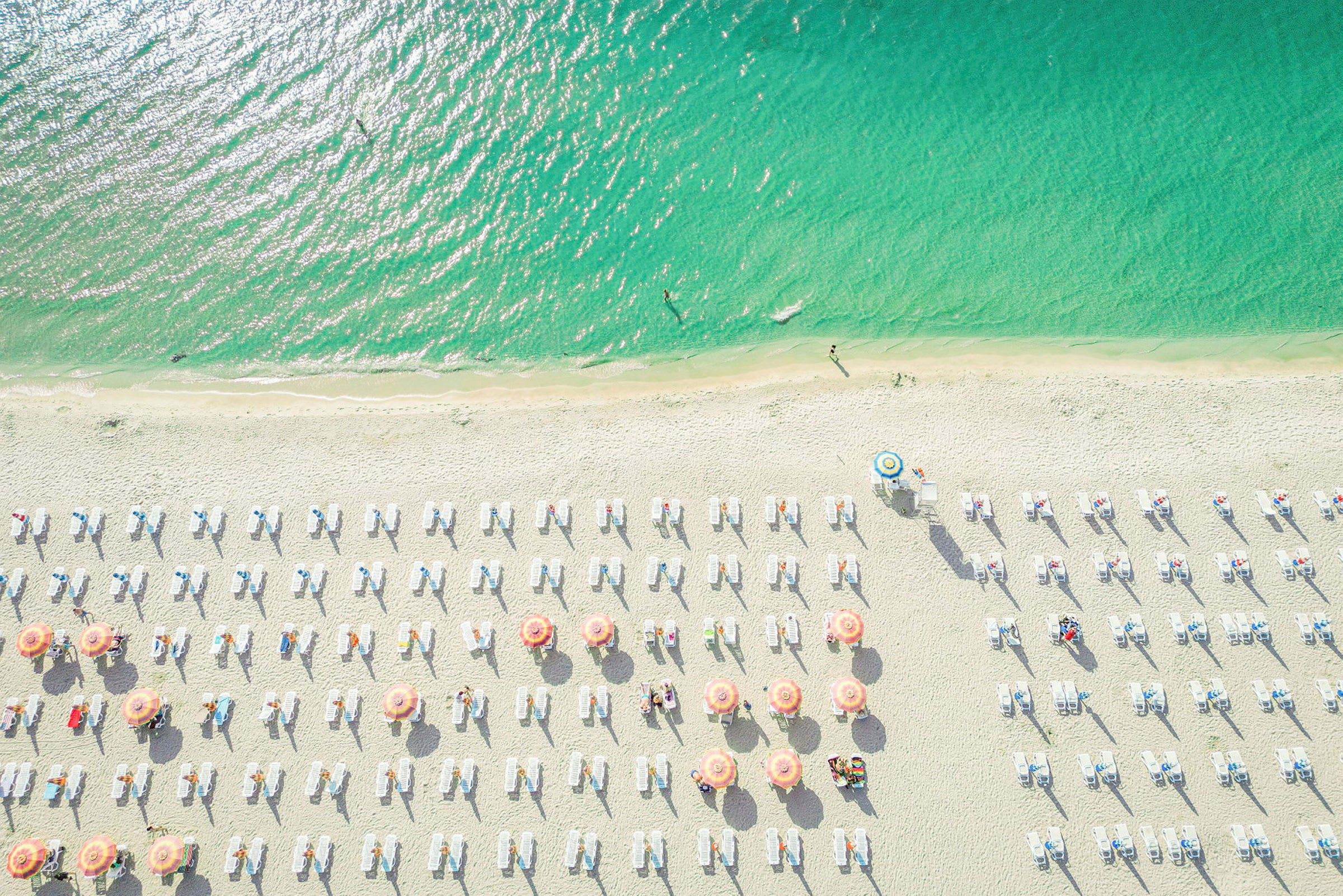 Flyfoto av en strand med parasoller og solsenger. Havet er tiltalende grønnblått. (AI-generert tekst)