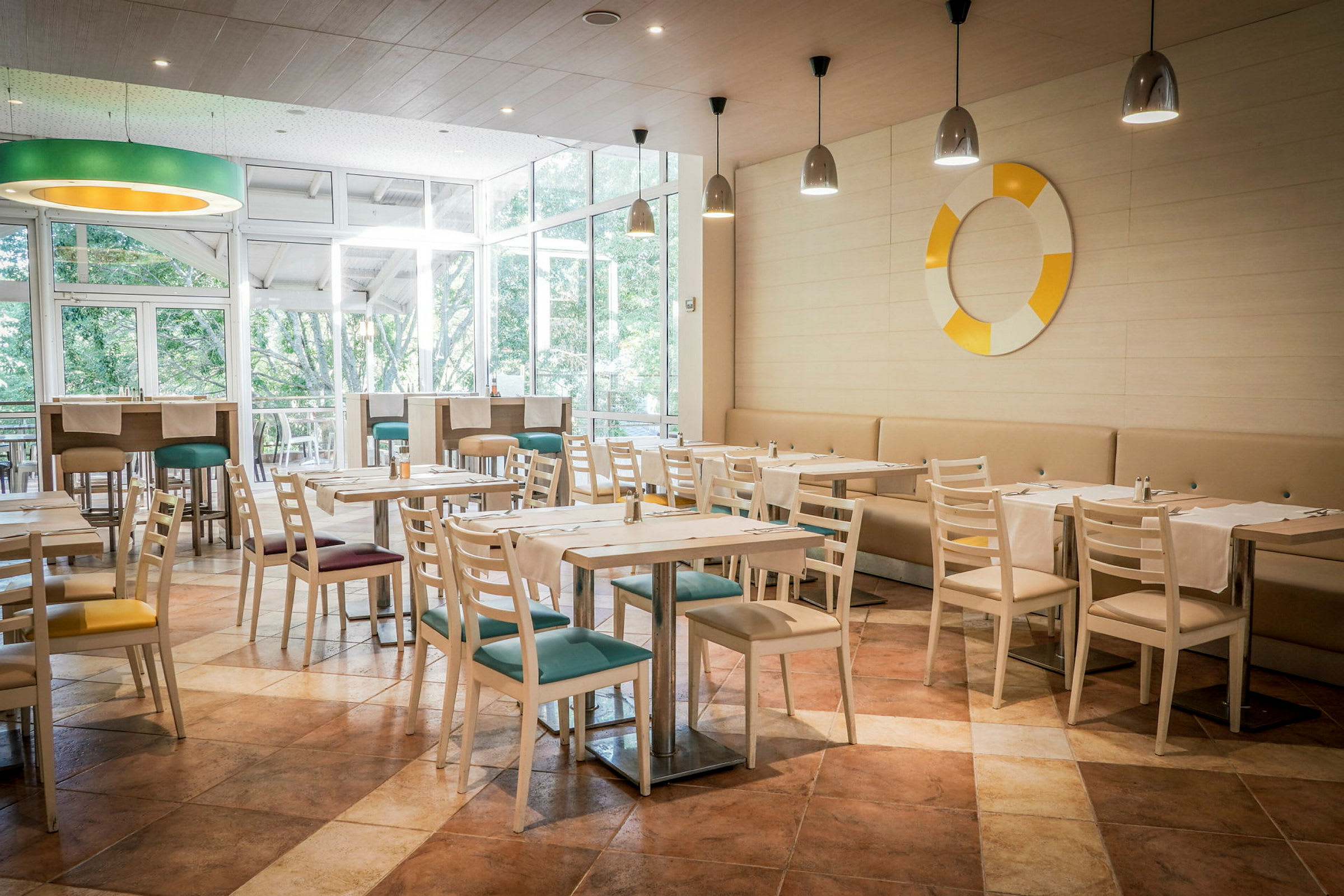 En restaurant med lyse trestoler og bord, fargerike sitteputer og vinduer mot grøntområder. (AI-generert tekst)