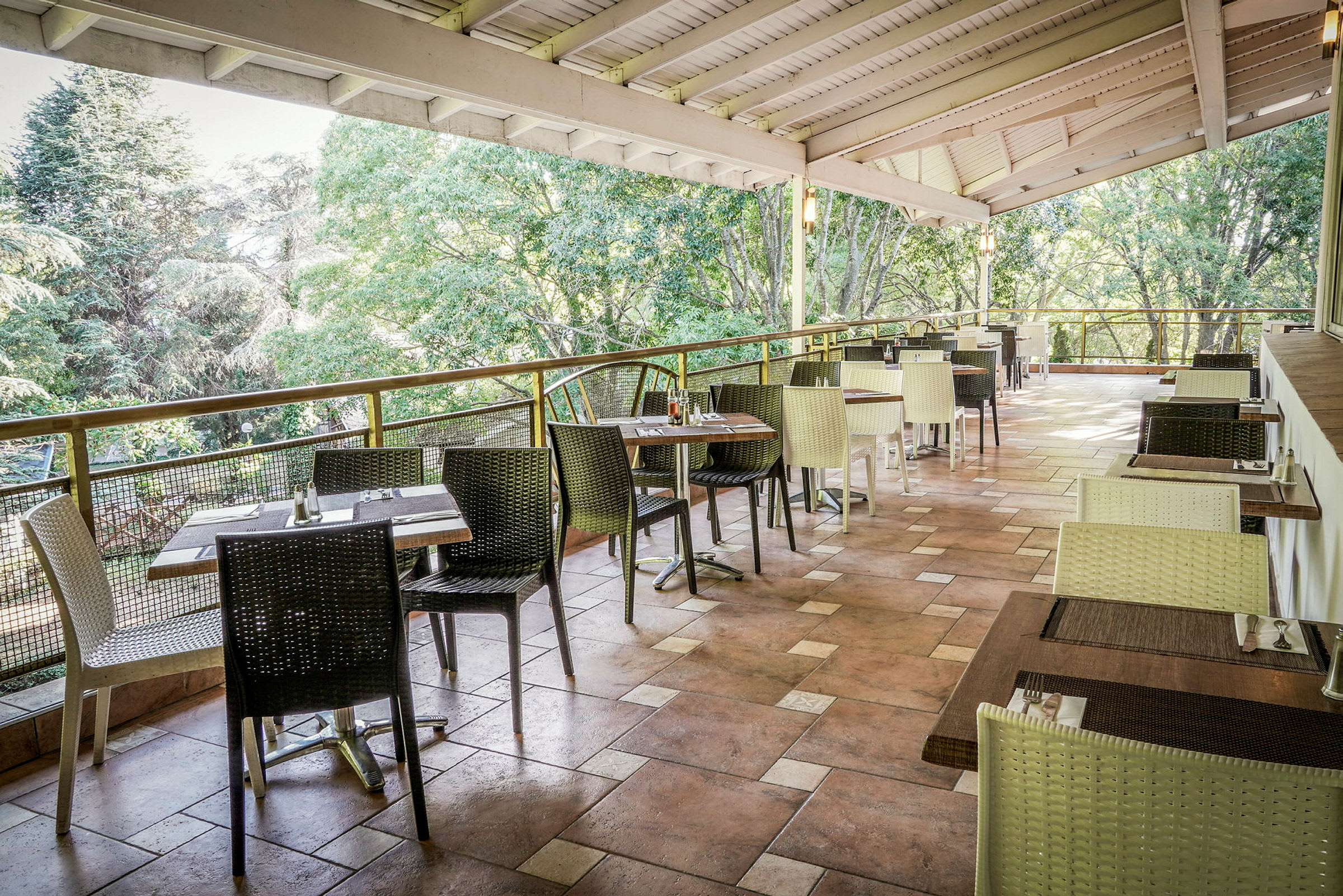 En åpen restaurantterrasse med bord og stoler, omgitt av grønne trær. (AI-generert tekst)