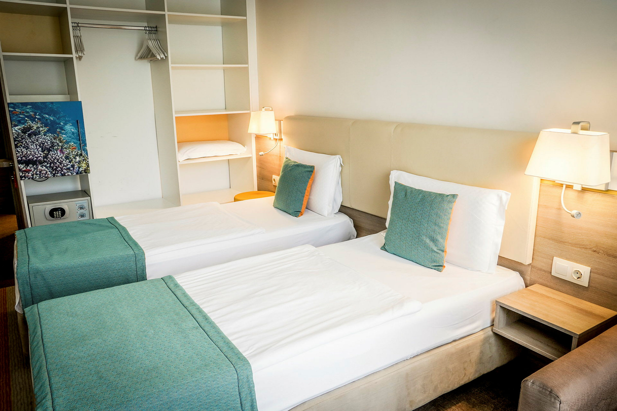 Hotellrom med to enkeltsenger, hvite laken og turkise puter. Innebygd oppbevaring og vegglamper vises. (AI-generert tekst)