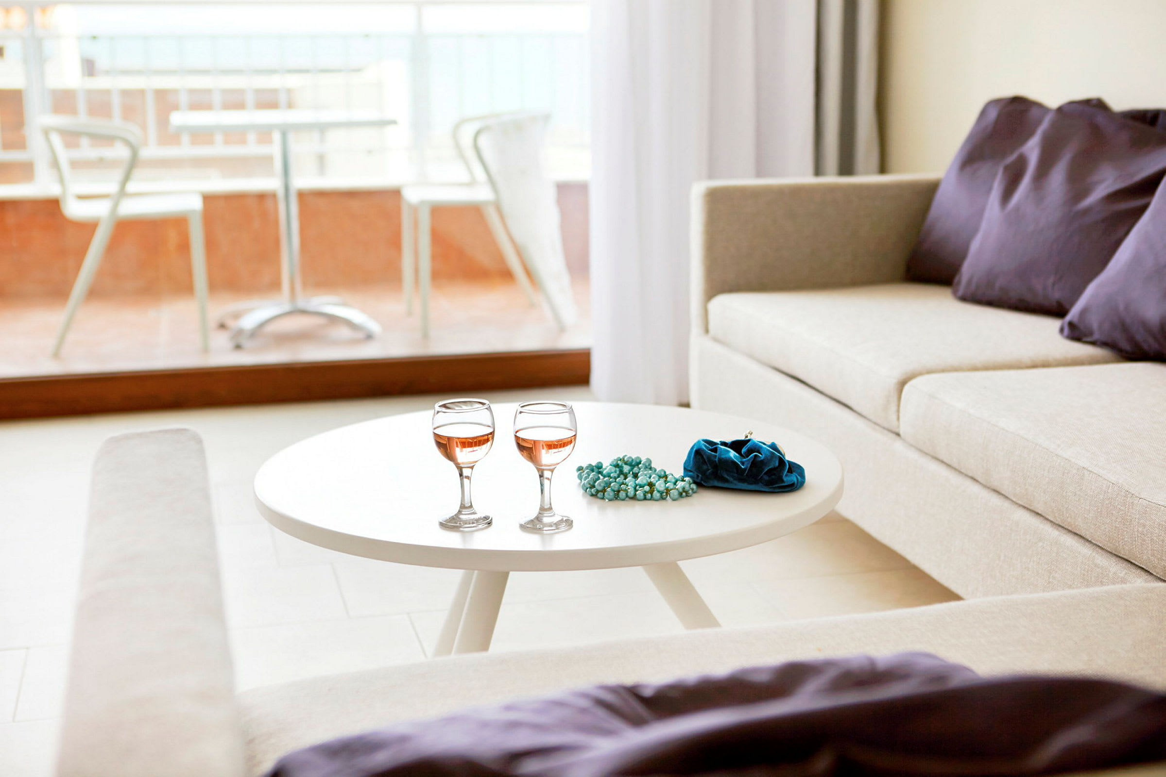 En lys stue med et hvitt sofabord som har to vinglass, et blått smykkesett eller pose og perler. En beige sofa med dekorative lilla puter ved siden av, og en balkong med bord og stoler synlig utenfor glassvinduet. (AI-generert tekst)