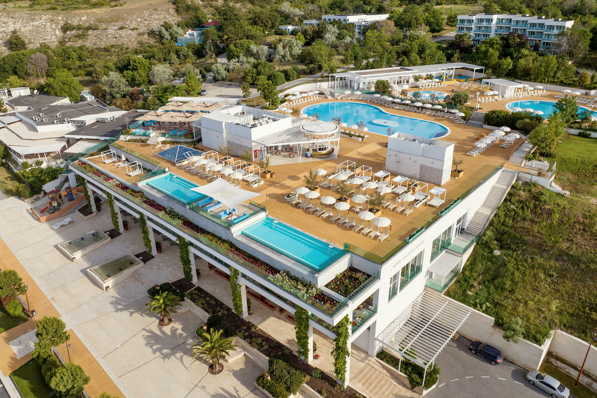 Et moderne hotellkompleks med flere utendørsbassenger, solsenger og parasoller, omgitt av grøntområder. (AI-generert tekst)