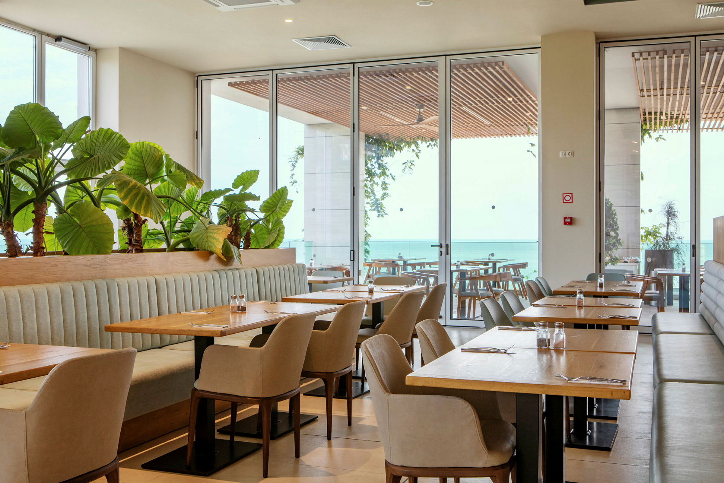 En restaurant med bord, stoler og planter ved store vinduer med utsikt over havet. (AI-generert tekst)