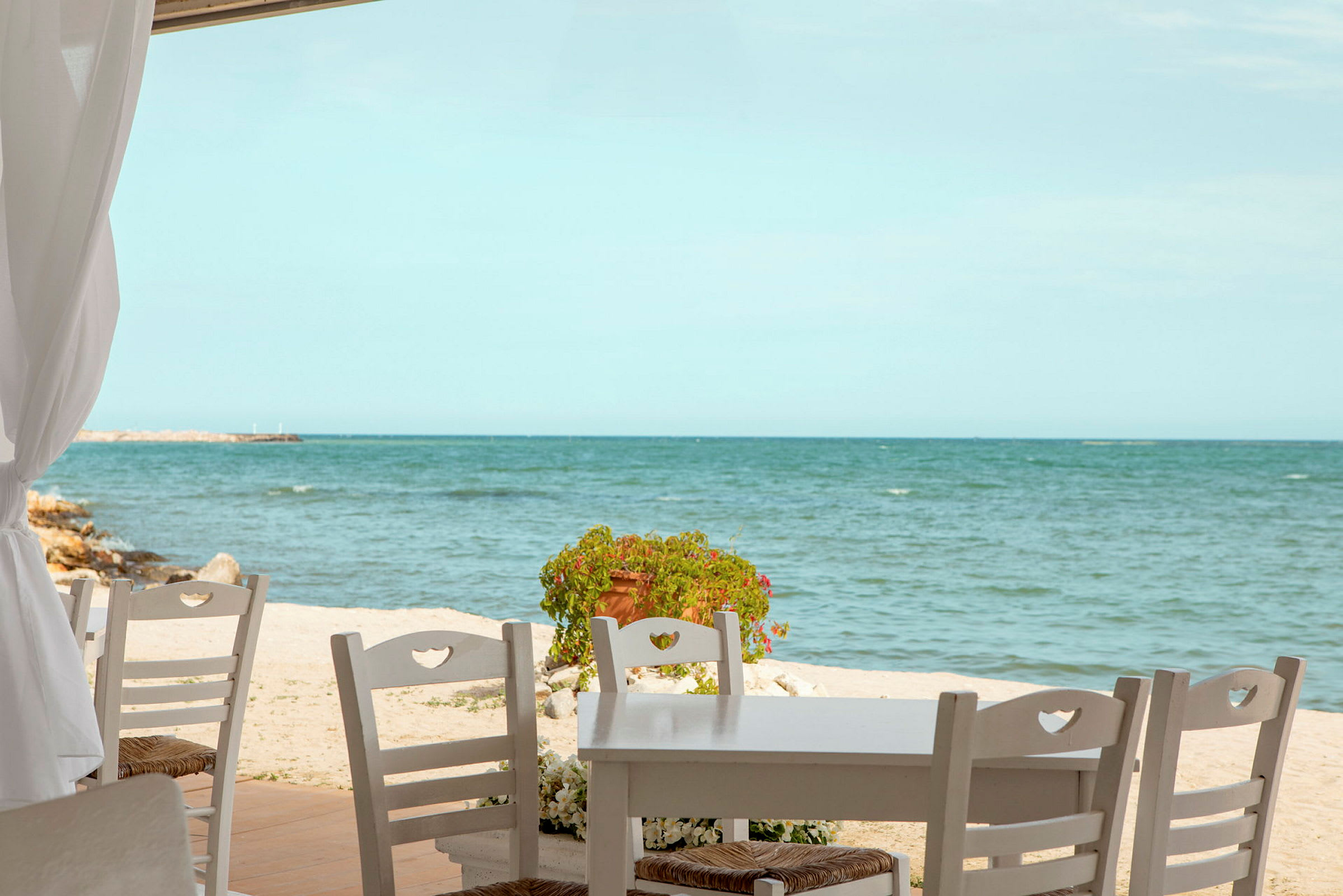 Et bord med trestoler står på en terrasse nær en strand med havet i bakgrunnen. (AI-generert tekst)