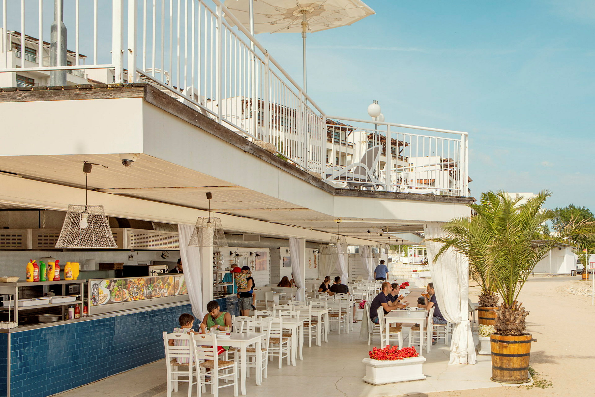 En utendørsrestaurant med hvite møbler, hengende lamper og en takterrasse. Folk sitter og spiser under taket. En palme er også synlig ved siden av restauranten. (AI-generert tekst)