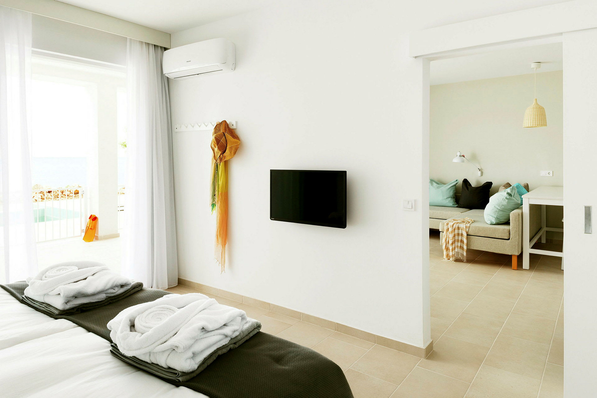 Et lyst hotellrom med en oppredd seng, en veggmontert TV og en sittegruppe. Balkong med utsikt kan sees i bakgrunnen. (AI-generert tekst)
