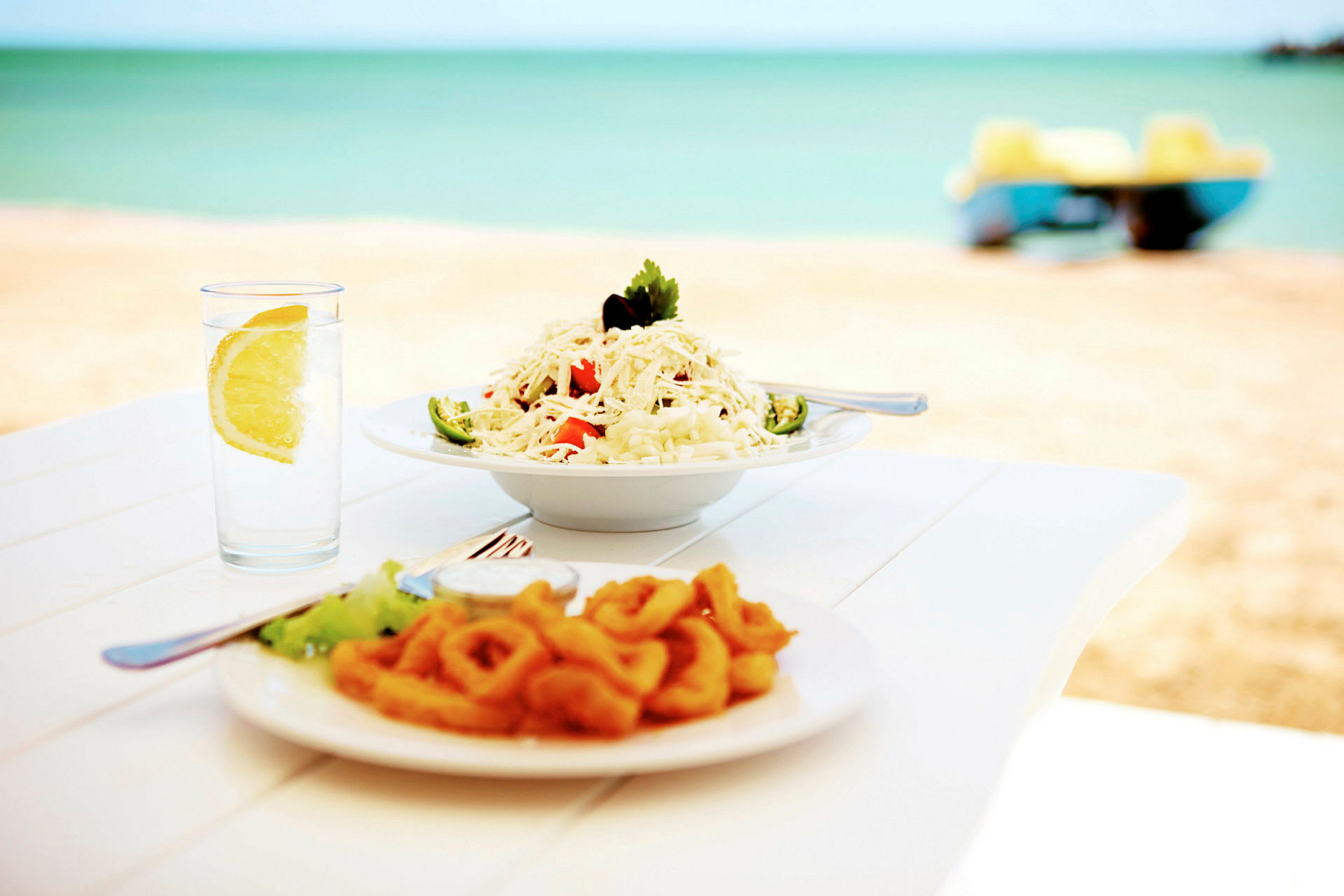 En tallerken med pasta og en tallerken med frityrstekte blekksprutringer servert ved et bord ved stranden, med et glass vann og en sitronskive ved siden av. (AI-generert tekst)