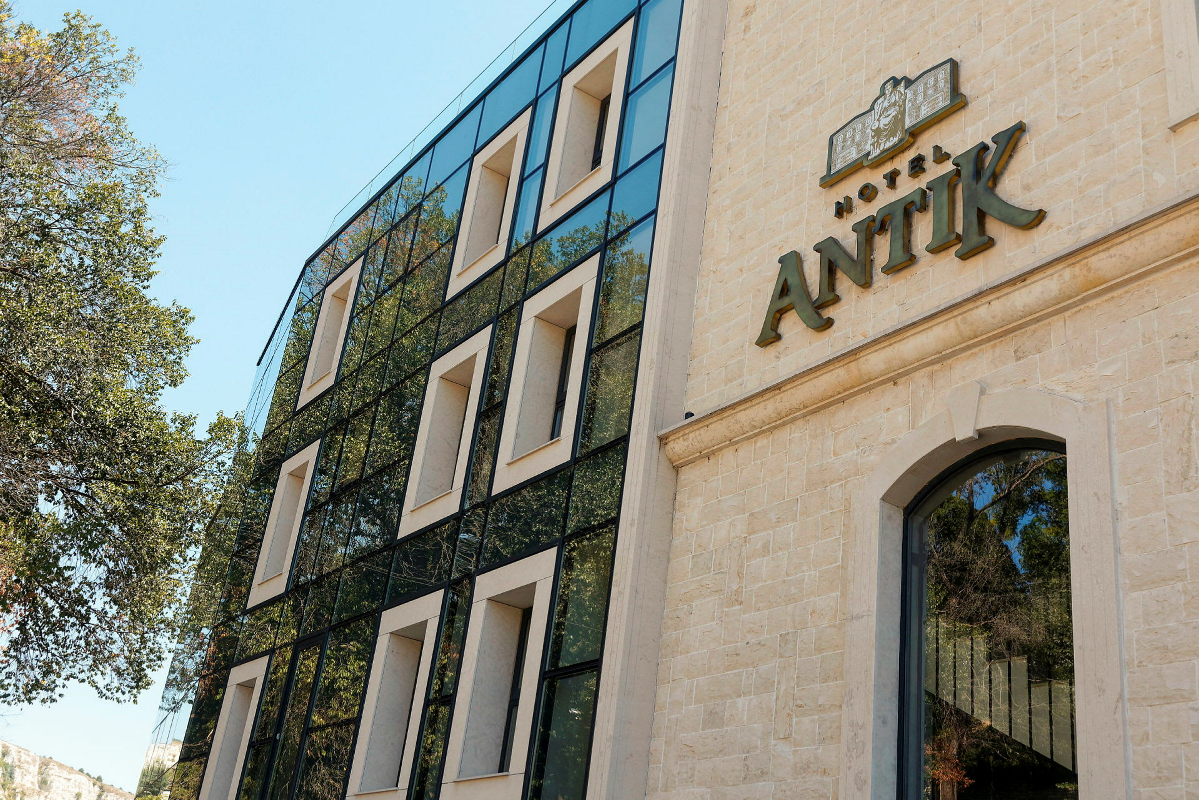Fasade av et hotell med navnet "Hotel Antik" synlig på bygningen. Den moderne bygningen har store vinduer og er omgitt av trær. (AI-generert tekst)