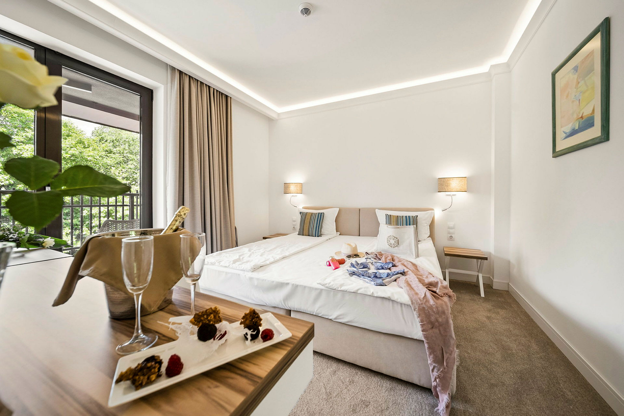 Hotellrom med dobbeltseng, nattbord og balkong med utsikt mot trær. Et bord med champagnekjøler og dessert. (AI-generert tekst)
