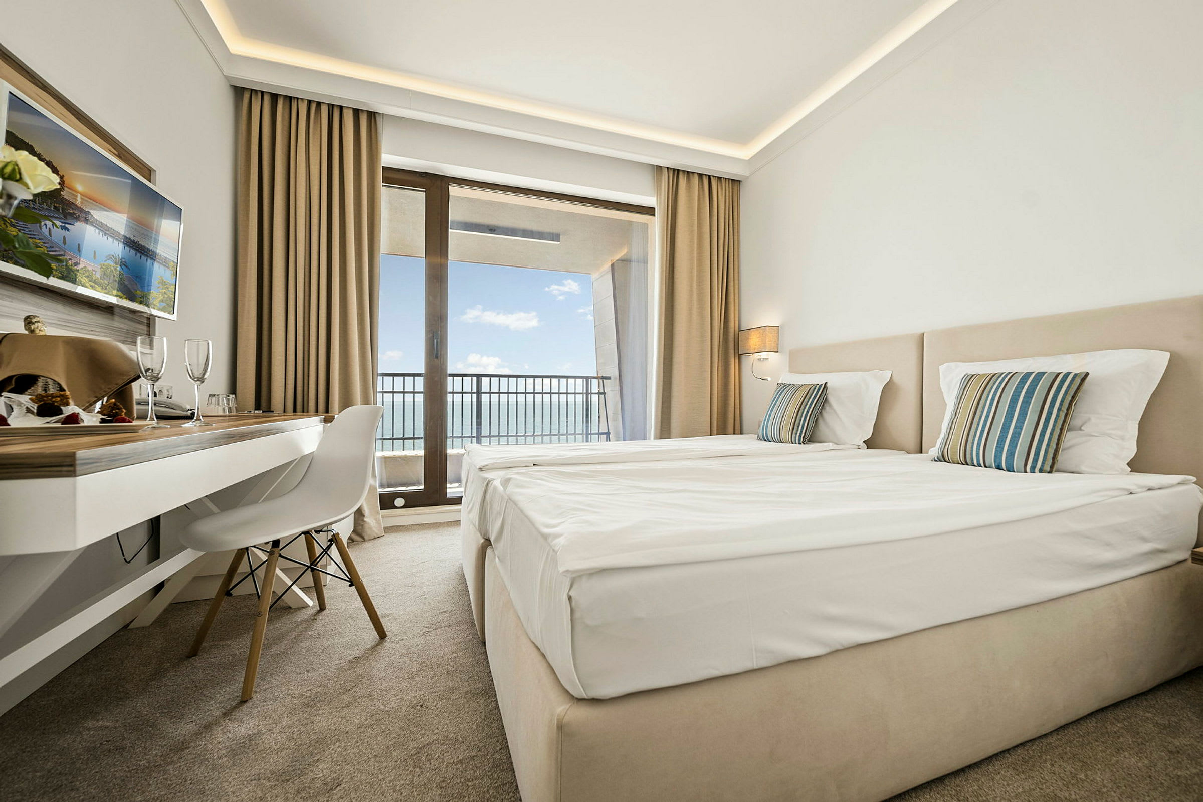 Hotellrom med to senger, skrivebord og balkong med utsikt over havet. (AI-generert tekst)