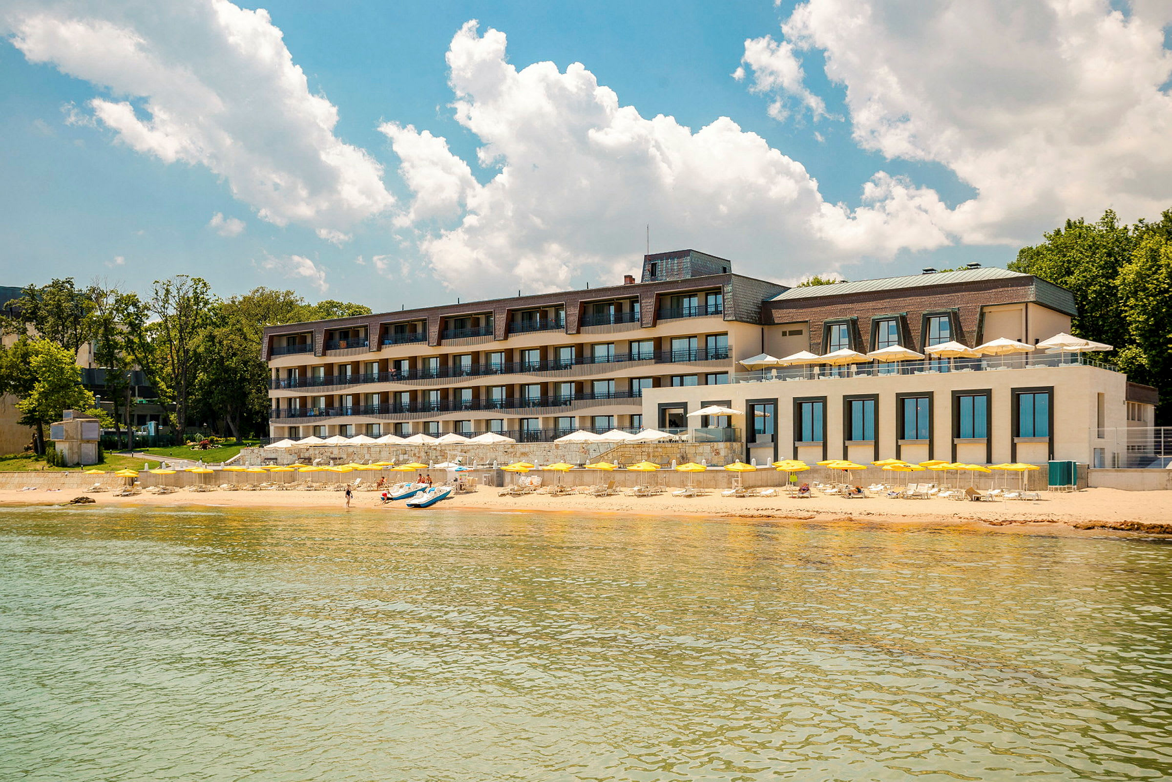 Fleretasjes hotell ved stranden med gule parasoller og klar blå himmel. (AI-generert tekst)