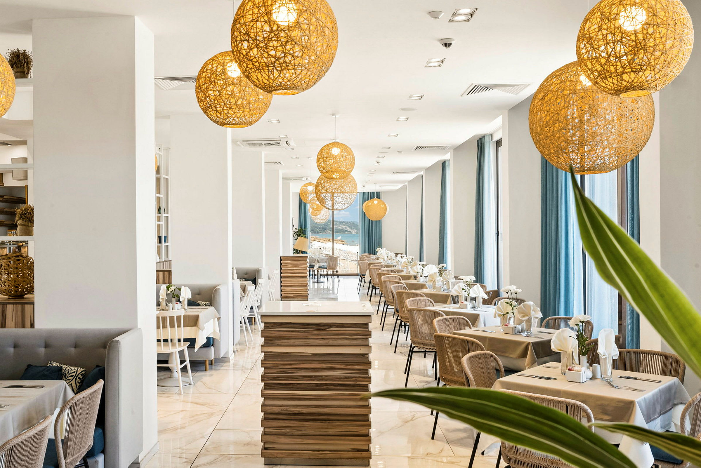 En moderne restaurant med en lys innredning som inkluderer trebord, stoler og store vinduer med blå gardiner. Dekorative belysningslamper henger fra taket. (AI-generert tekst)