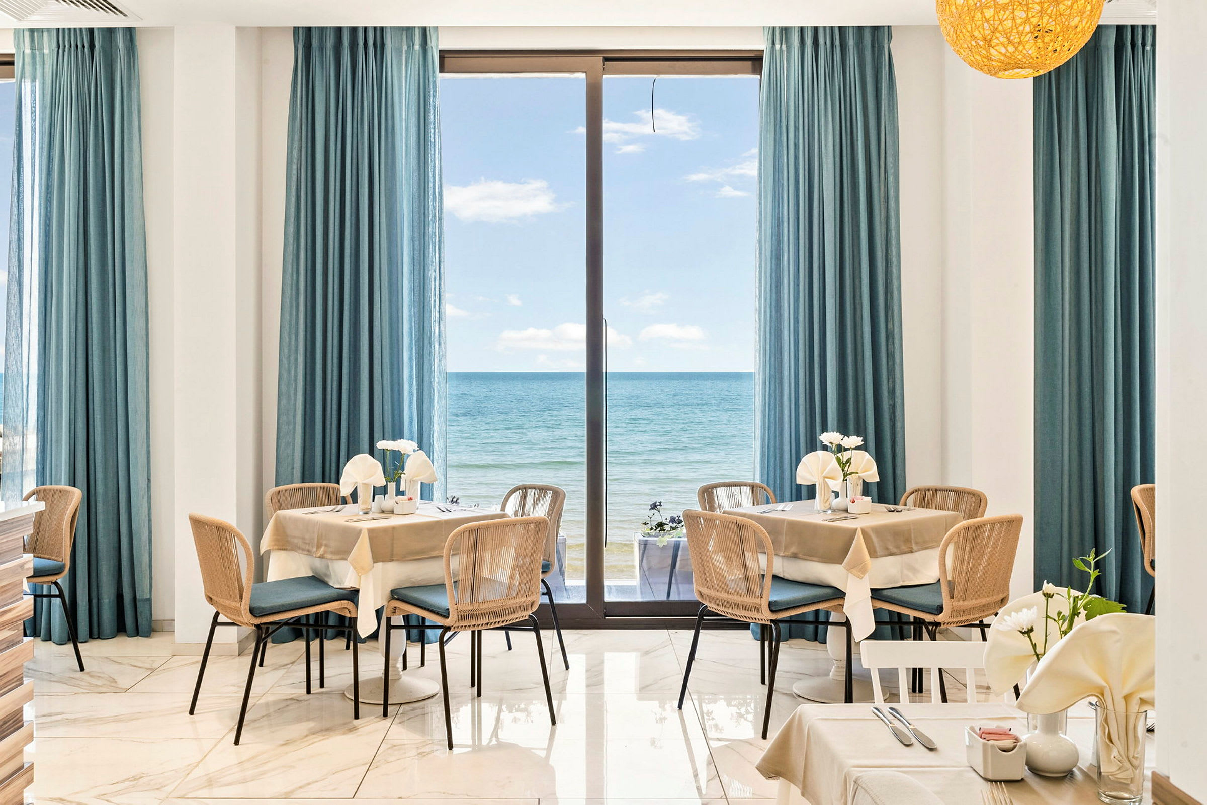 Interiør av en restaurant med bord og stoler, vinduer dekket med blå gardiner, og utsikt over havet. (AI-generert tekst)