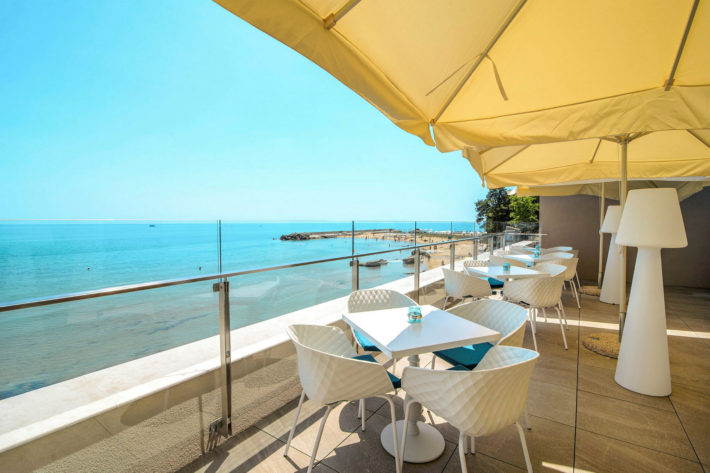 Terrasse med bord og stoler under parasoller som vender ut mot stranden og havet. (AI-generert tekst)