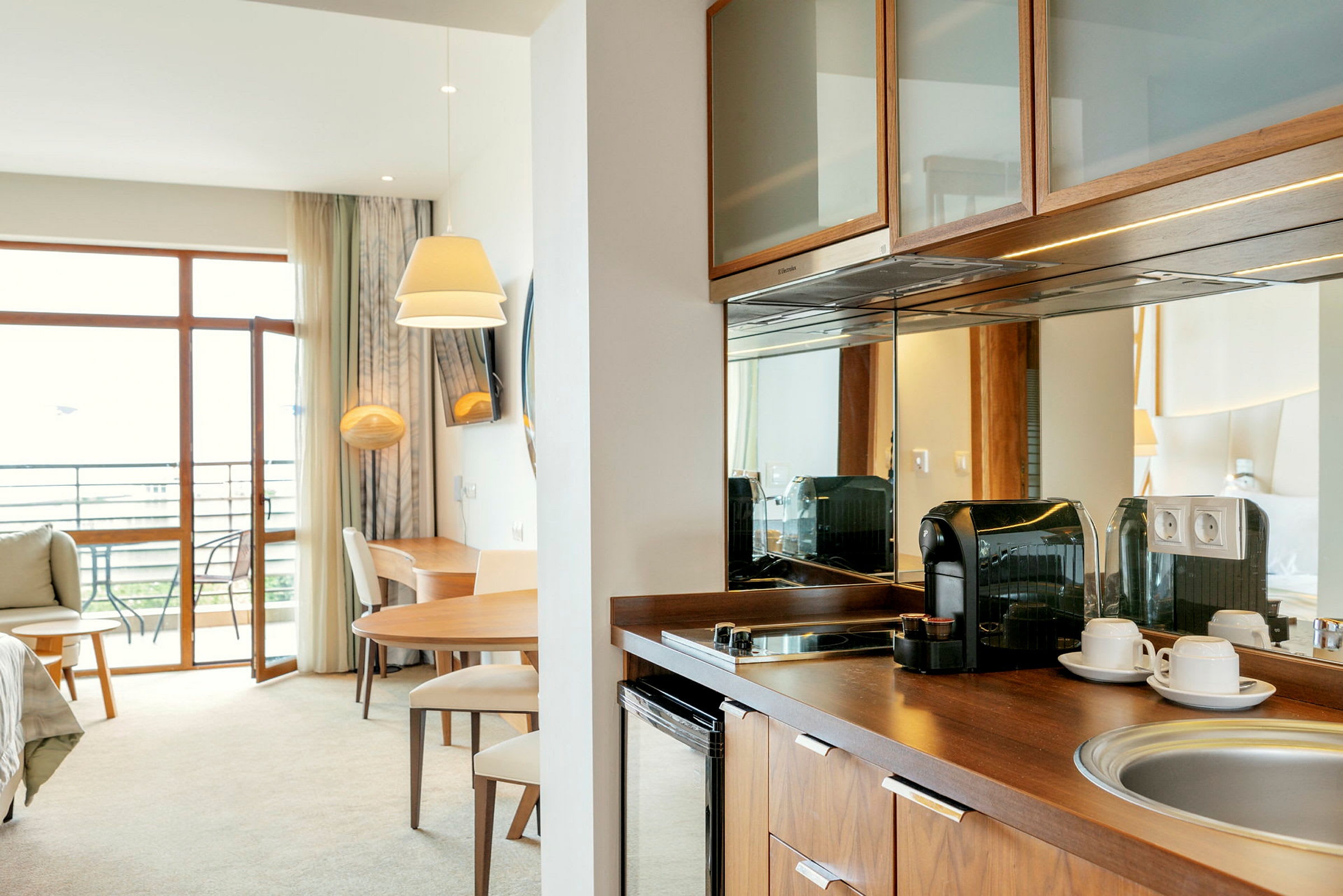 Moderne hotellrom med balkong, bord, stoler, kaffetrakter og kjøkkenkrok. (AI-generert tekst)