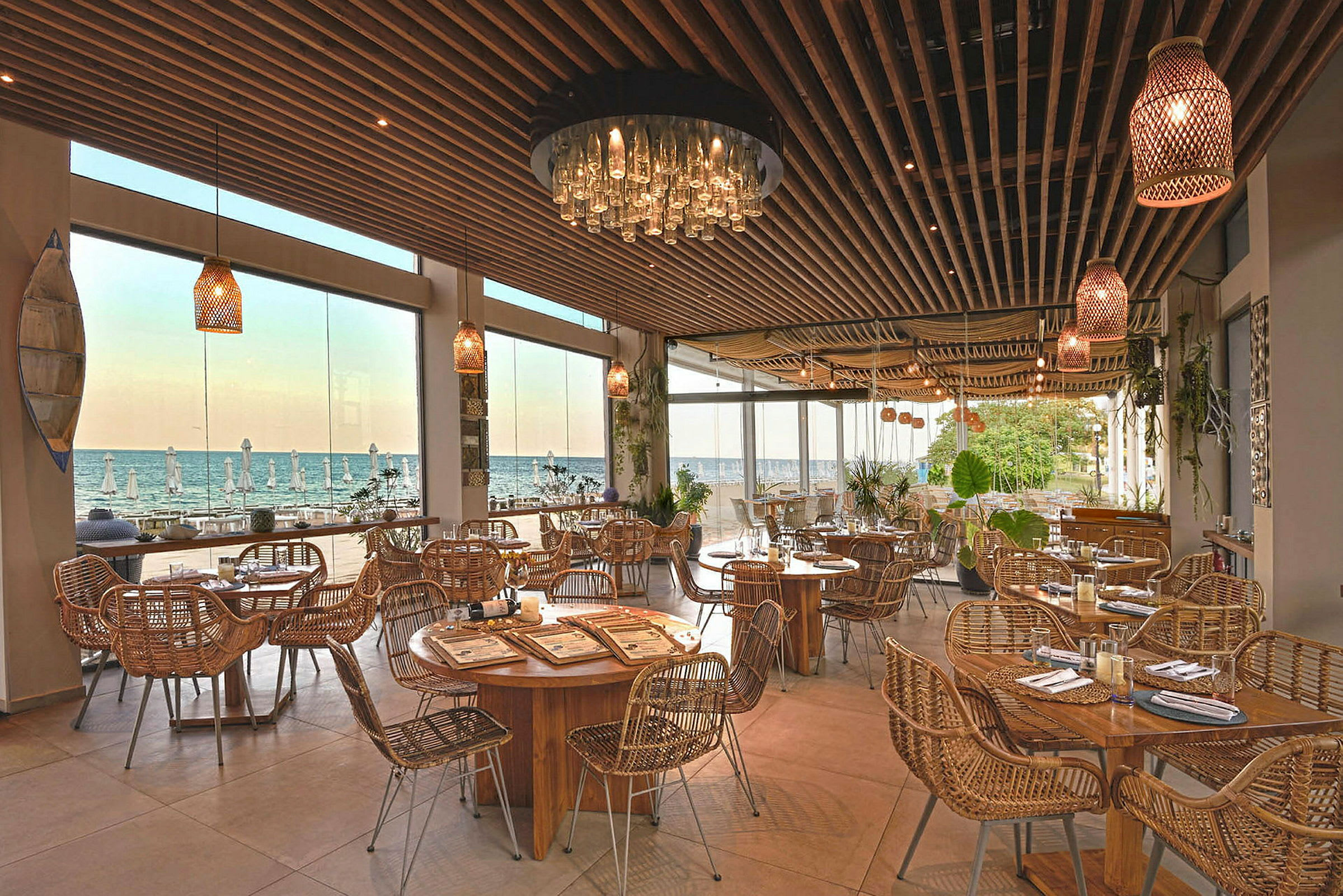 Restaurant med trebord og rottingstoler, store vinduer viser utsikt mot havet. (AI-generert tekst)