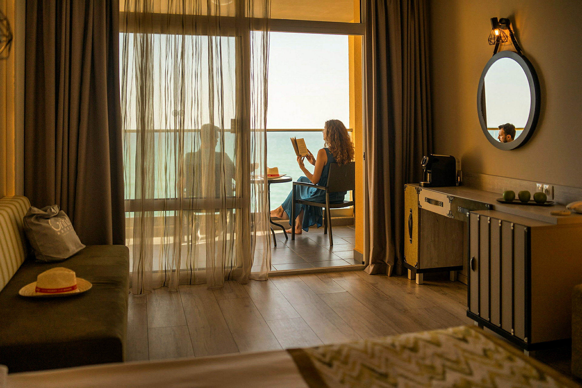 Hotellrom med en balkong der noen sitter og leser med utsikt over havet. (AI-generert tekst)