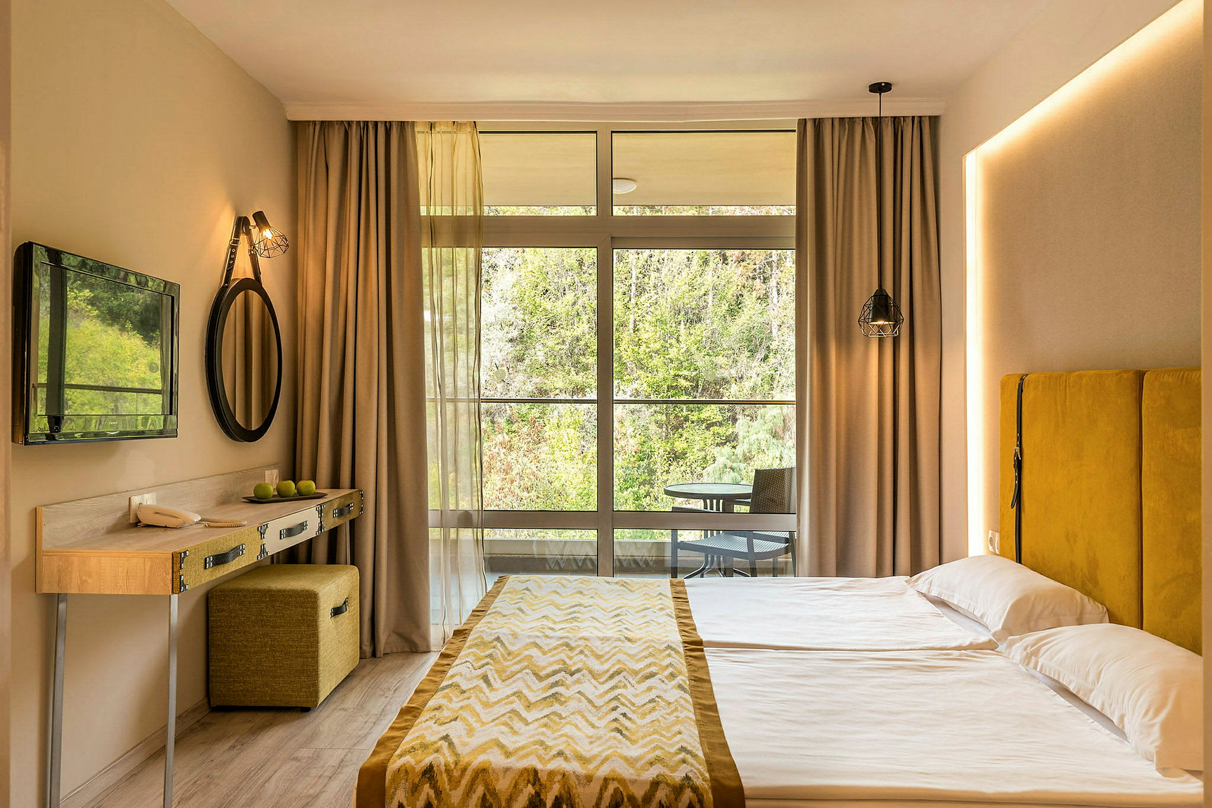 Et hotellrom med en dobbeltseng, gul sengegavl, lite skrivebord, og en stor vindusfront mot naturen. (AI-generert tekst)