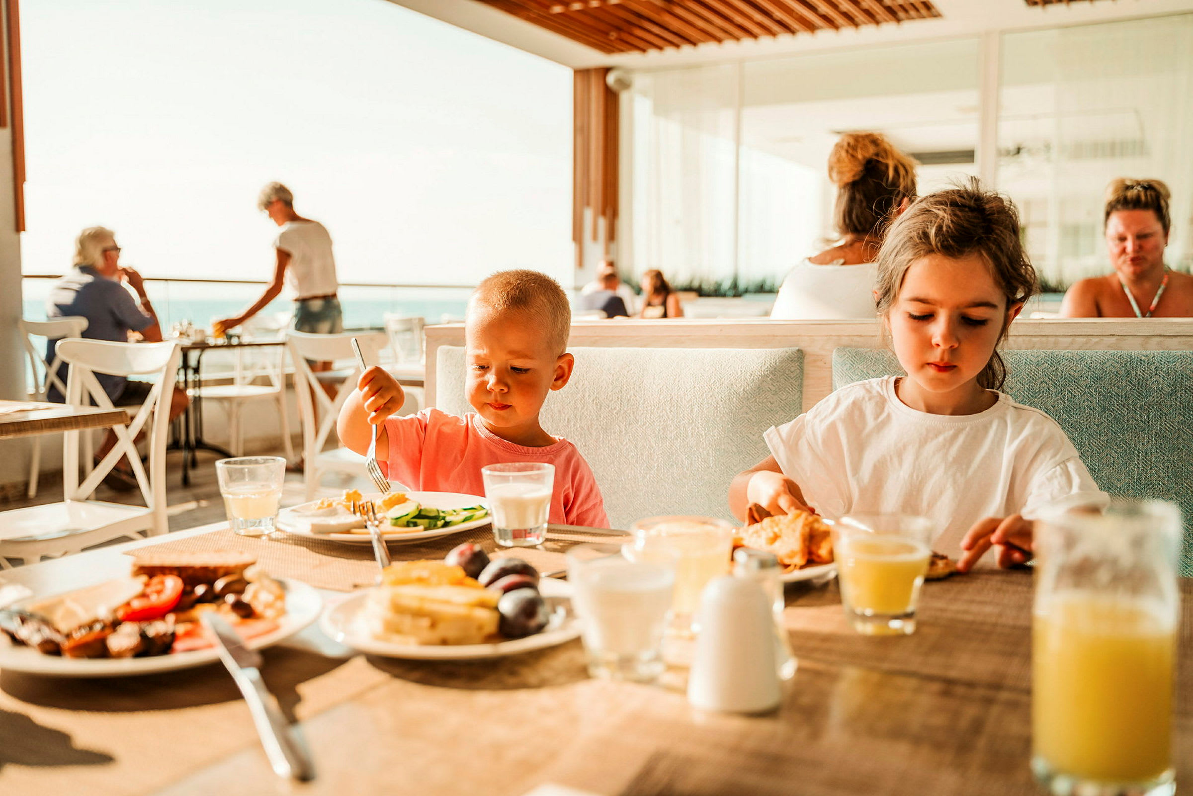 Barn sitter ved et bord og spiser mens mennesker ses i bakgrunnen på en restaurant med utsikt. (AI-generert tekst)