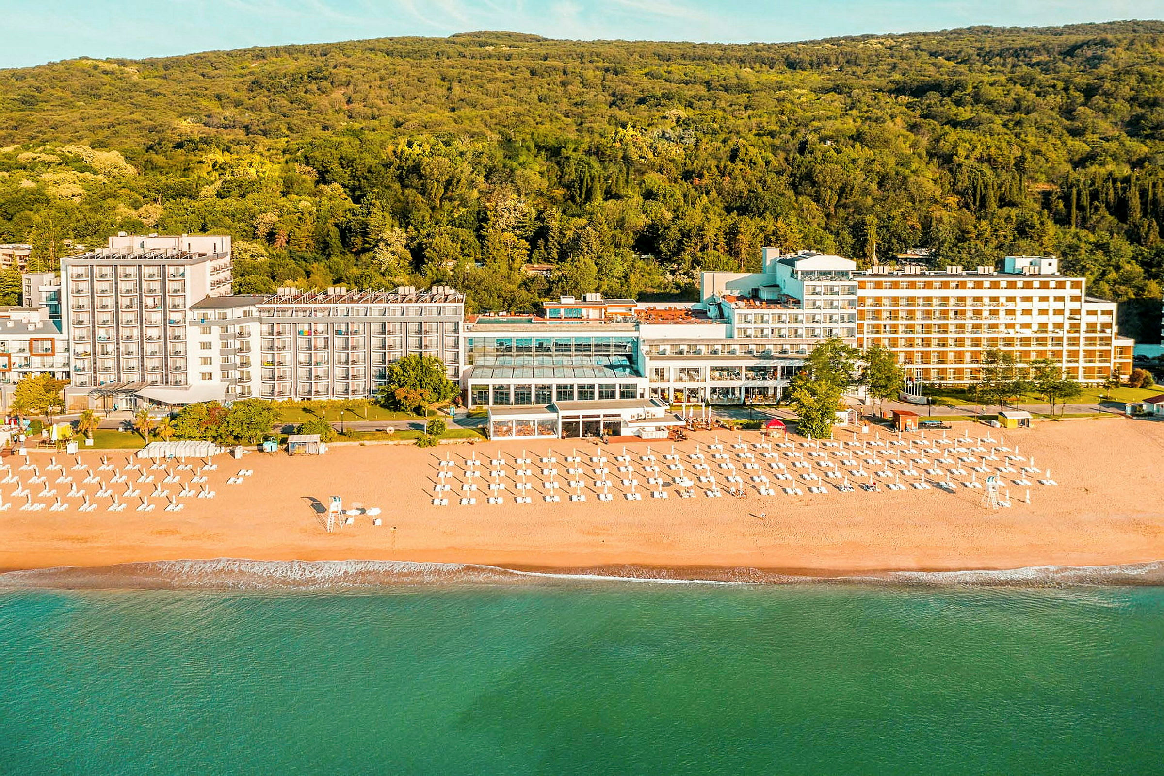 Hotell ved stranden med solsenger og parasoller foran en skog. (AI-generert tekst)