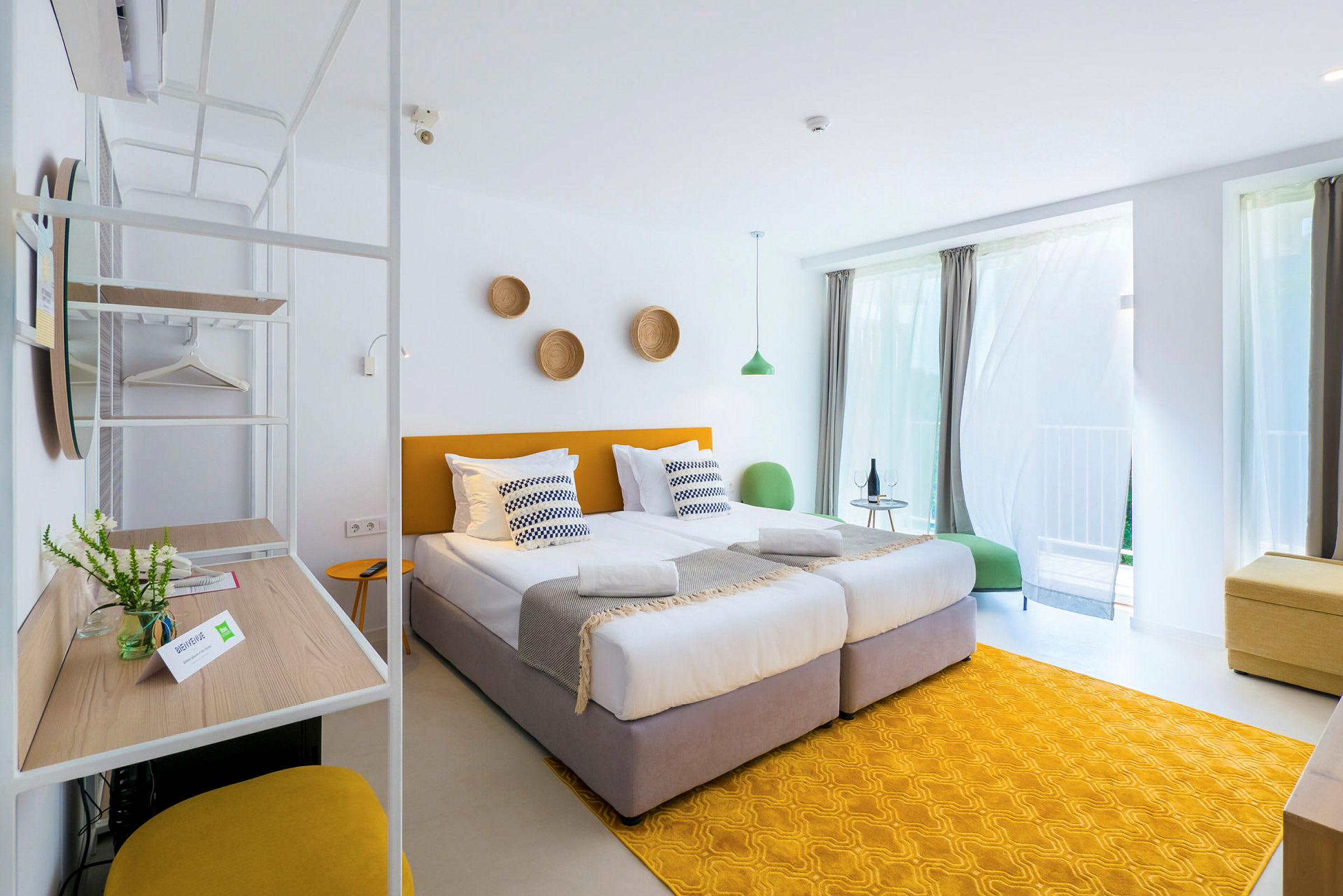Et lyst og moderne hotellrom med to enkeltsenger, gule detaljer og store vinduer med gardiner. (AI-generert tekst)