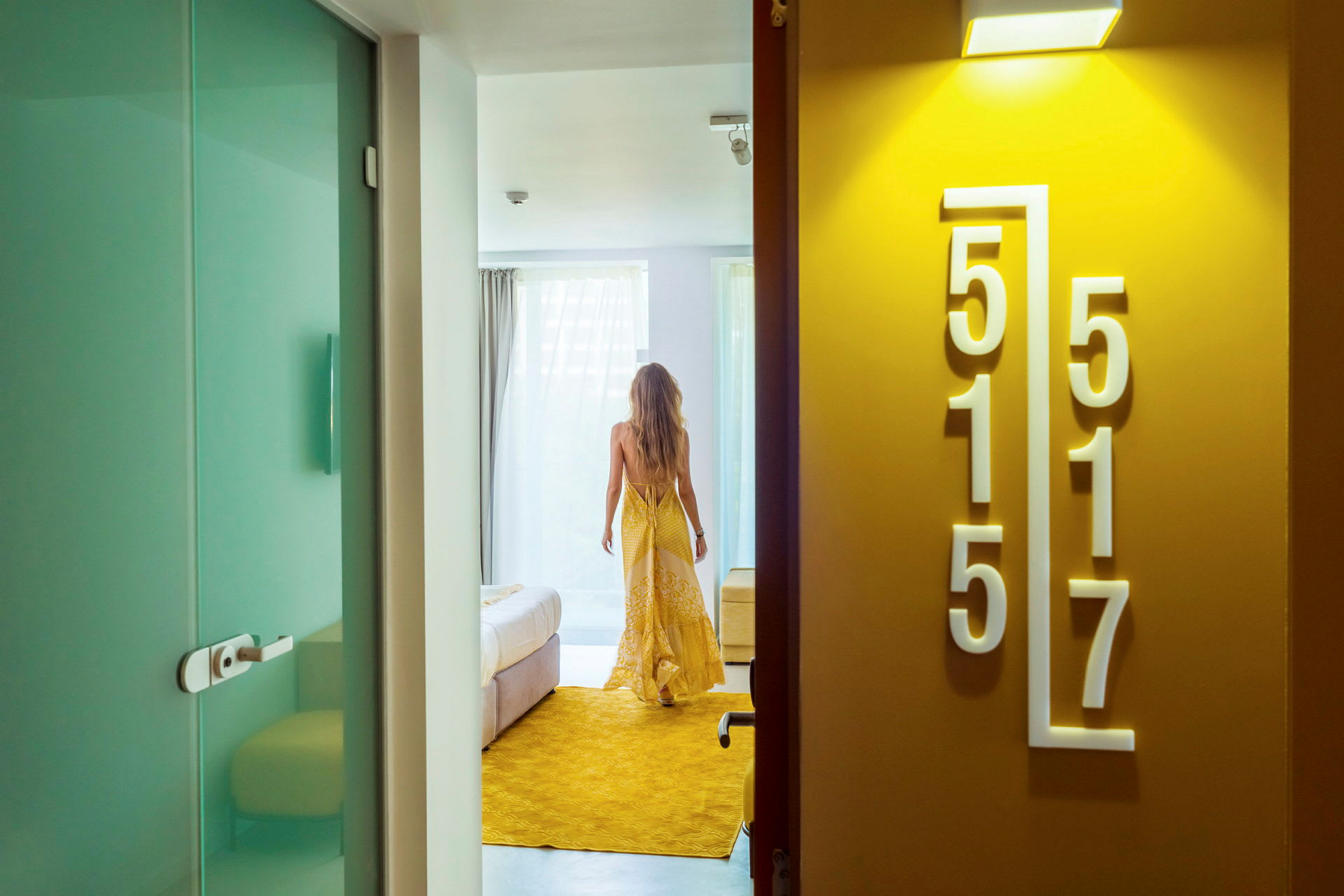En person i en gul kjole som går inn på et hotellrom med lys innredning. (AI-generert tekst)