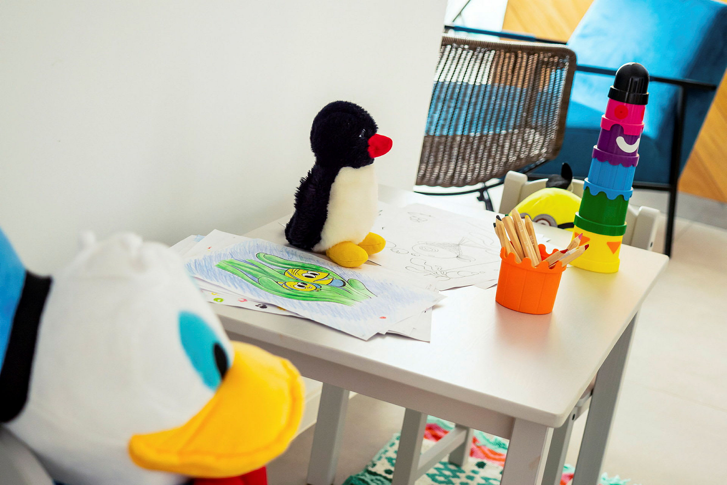 Et bord med tegninger, blyanter, en lekepinguin og fargerike stableleker. (AI-generert tekst)
