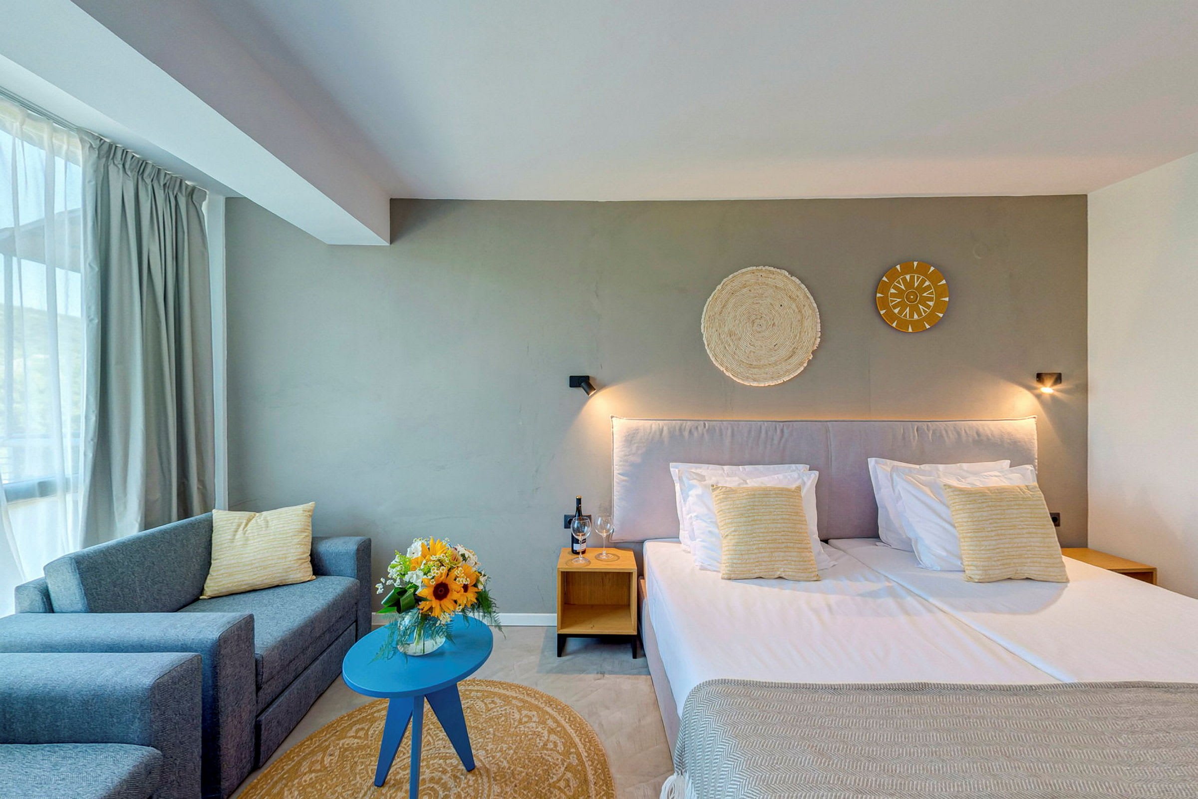 Et lyst innredet hotellrom med en dobbeltseng, sofa og et lite bord. På bordet står en vase med blomster. (AI-generert tekst)