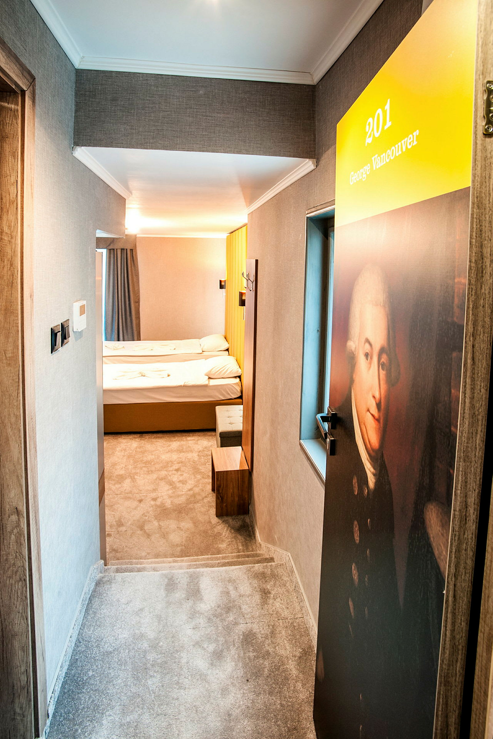 En hotellkorridor med en dør merket med nummer 201 og navnet George Vancouver. Innenfor kan man se et enkelt hotellrom med to senger. (AI-generert tekst)