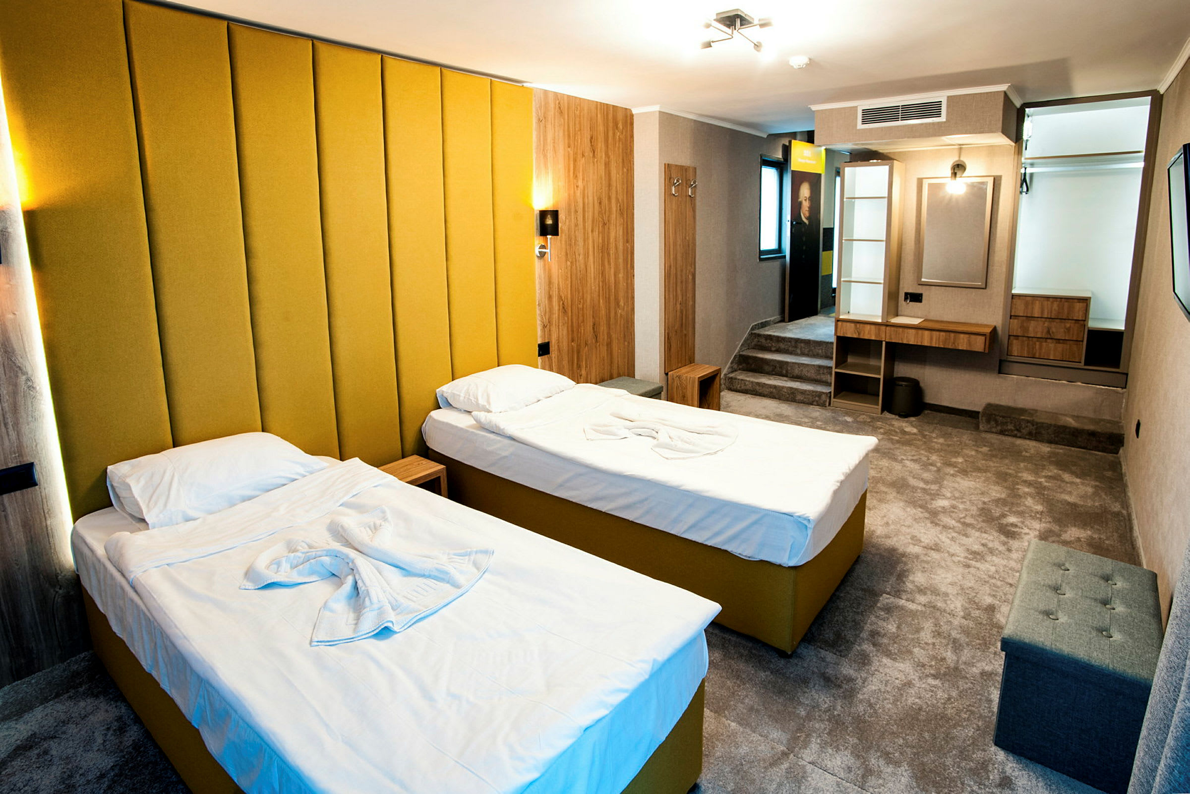 Et hotellrom med to enkeltsenger med gul polstring og hvitt sengetøy. Rommet har en moderne innredning med en gul veggpanel, et skrivebord, sittemøbler og en liten oppbevaringshylle. (AI-generert tekst)