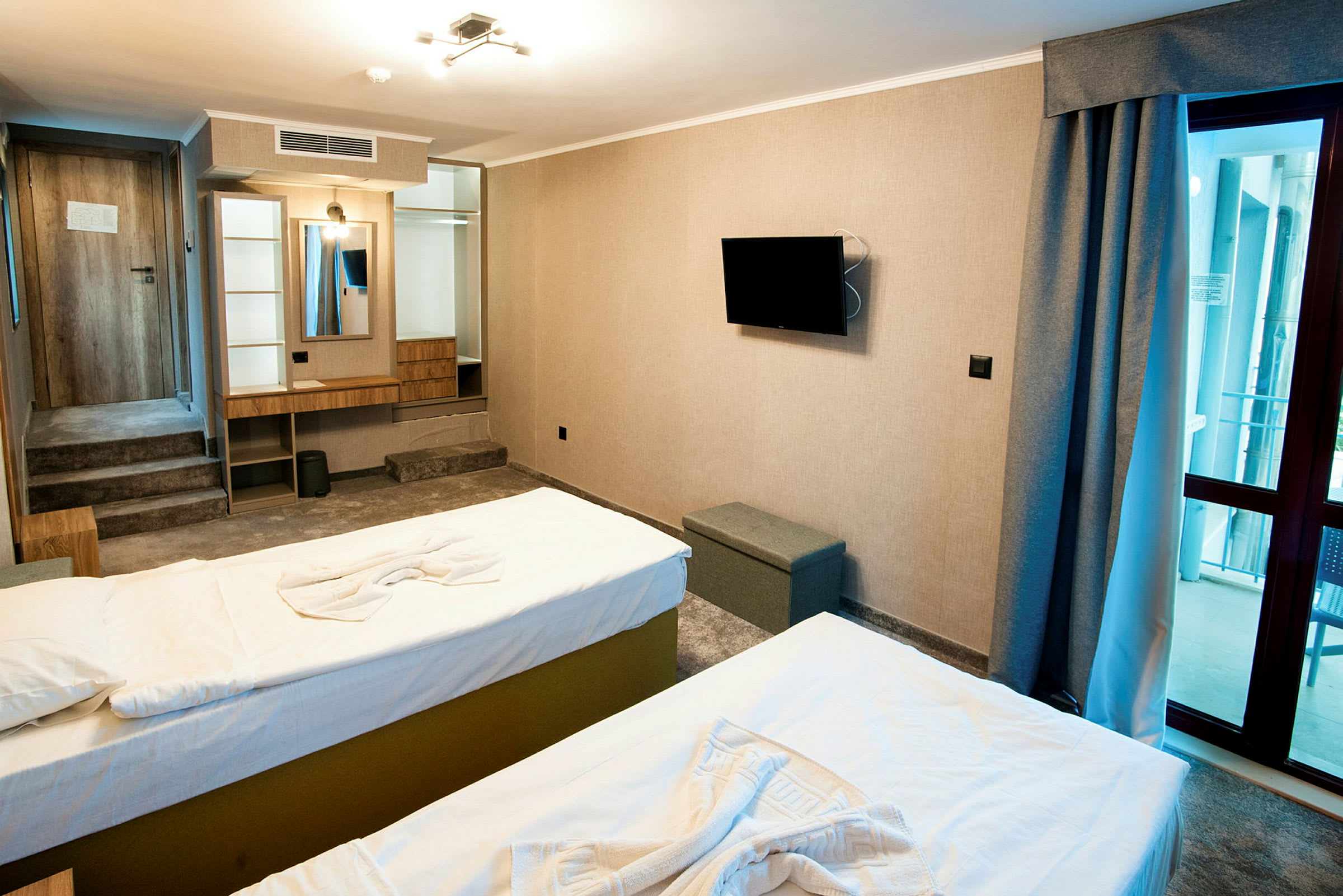 Et hotellrom med to senger, en veggmontert TV, speil med skrivebord og en glassdør mot balkong. (AI-generert tekst)